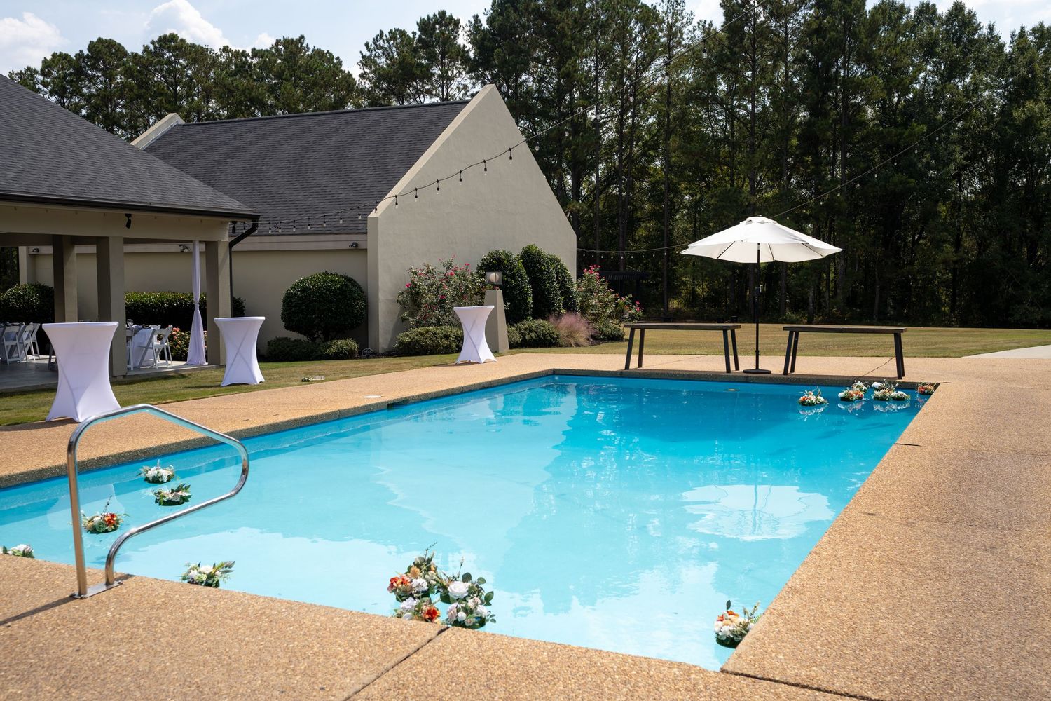 Swimming pool area with white umbrellas and lounge chairs surrounded by trees on a sunny day.