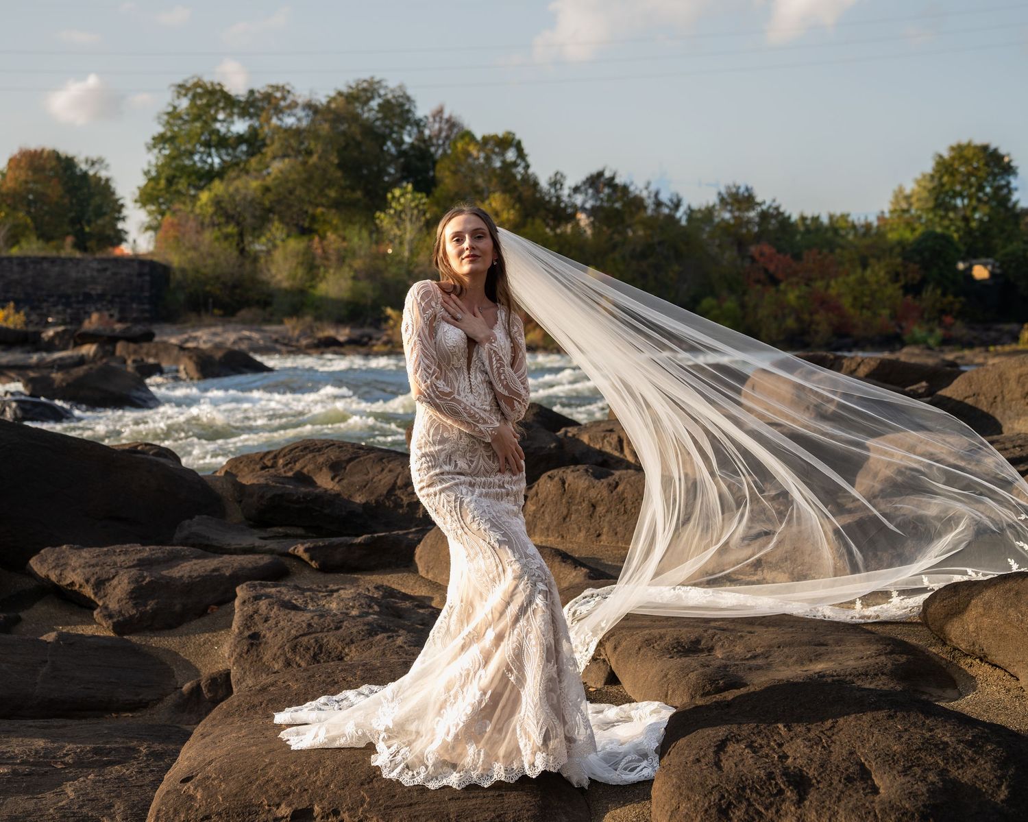 Scenic bridal portrait on rocky shoreline with flowing veil and autumn foliage.