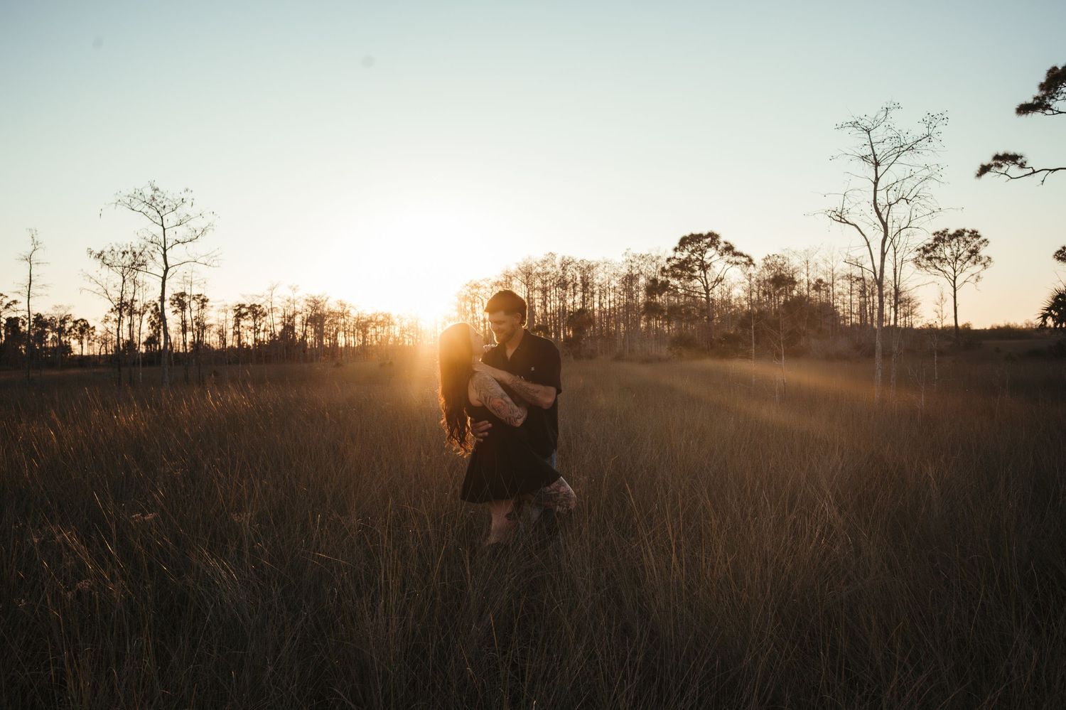 A silhouette stands in a golden field at sunset with tall trees in the background.