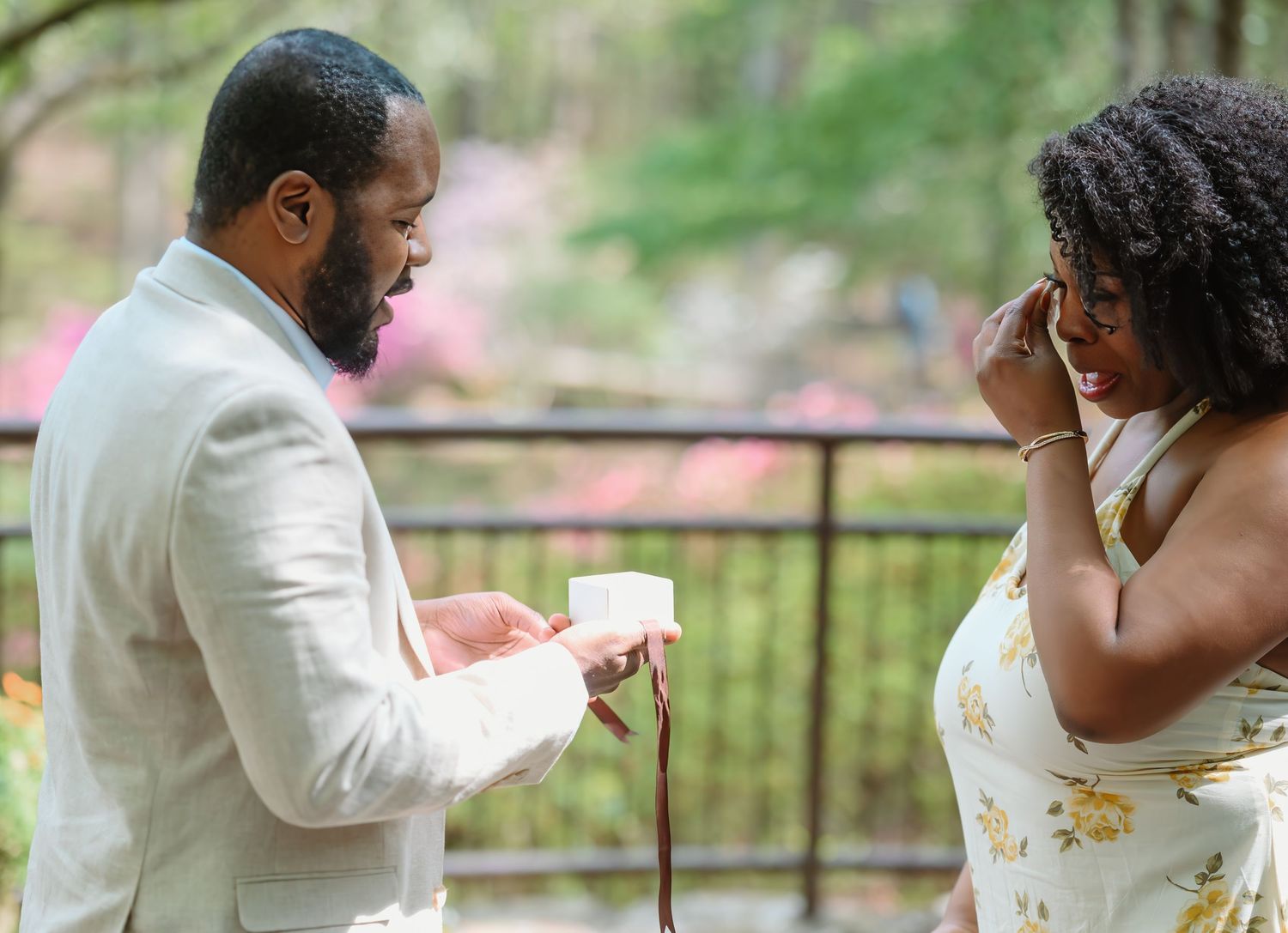 Man in white suit opens ring box while emotional woman wipes tears during outdoor marriage proposal.