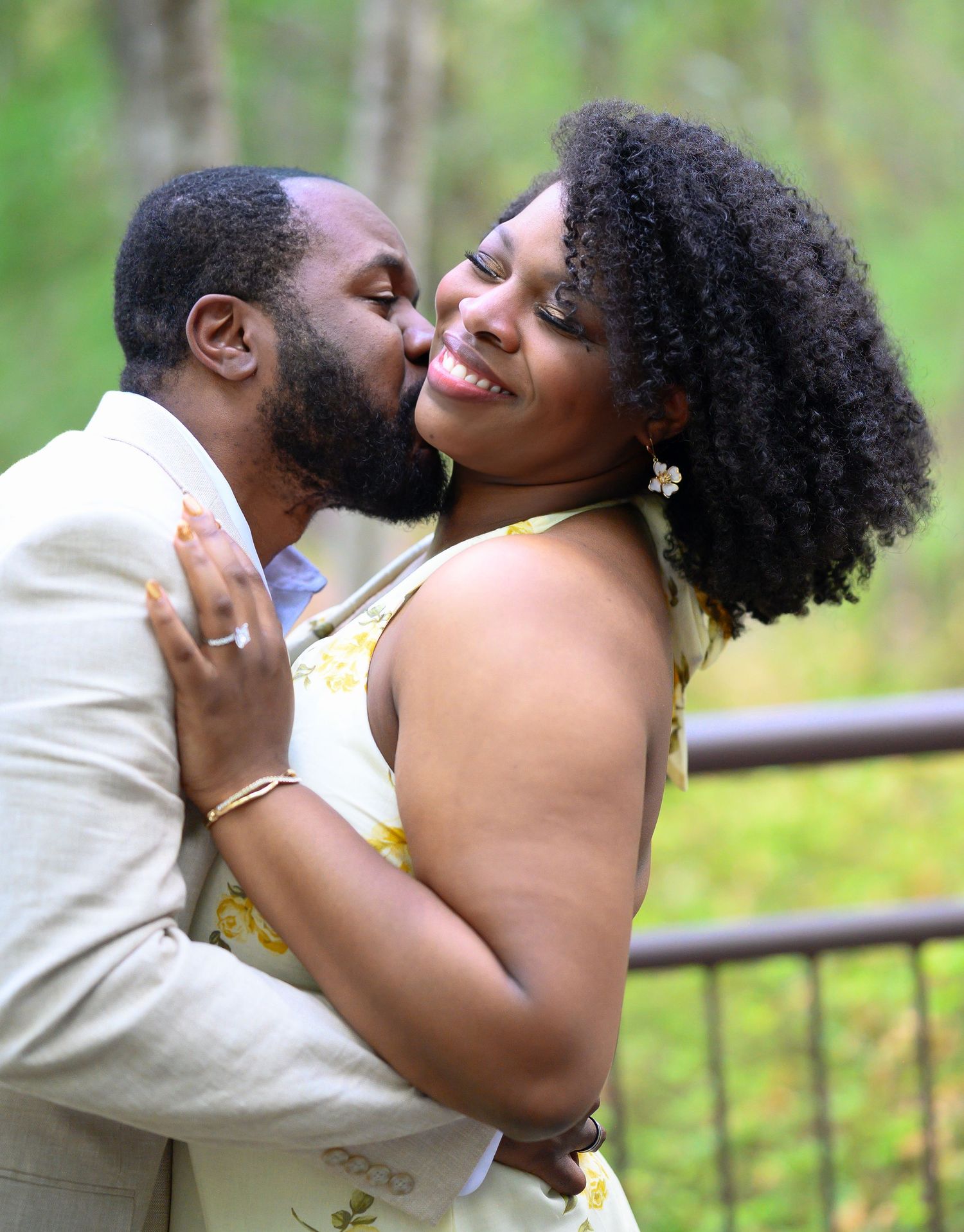 Man kissing woman on the cheek as she smiles joyfully outdoors in a lush green garden setting.
