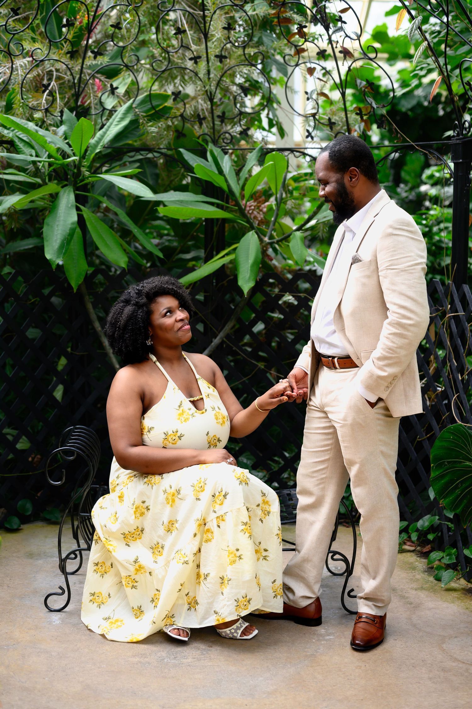 Man in beige suit holds hands with woman in yellow floral dress seated on ornate bench in tropical garden.
