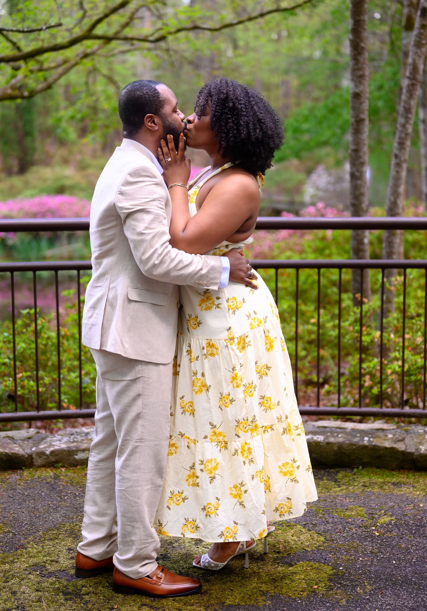 A couple shares a passionate kiss in a lush garden with pink azaleas, man in beige suit and woman in yellow floral dress.