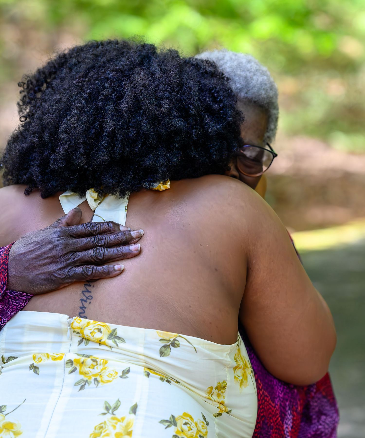 Older woman with gray hair embracing young woman in yellow floral dress in a warm congratulatory hug outdoors.