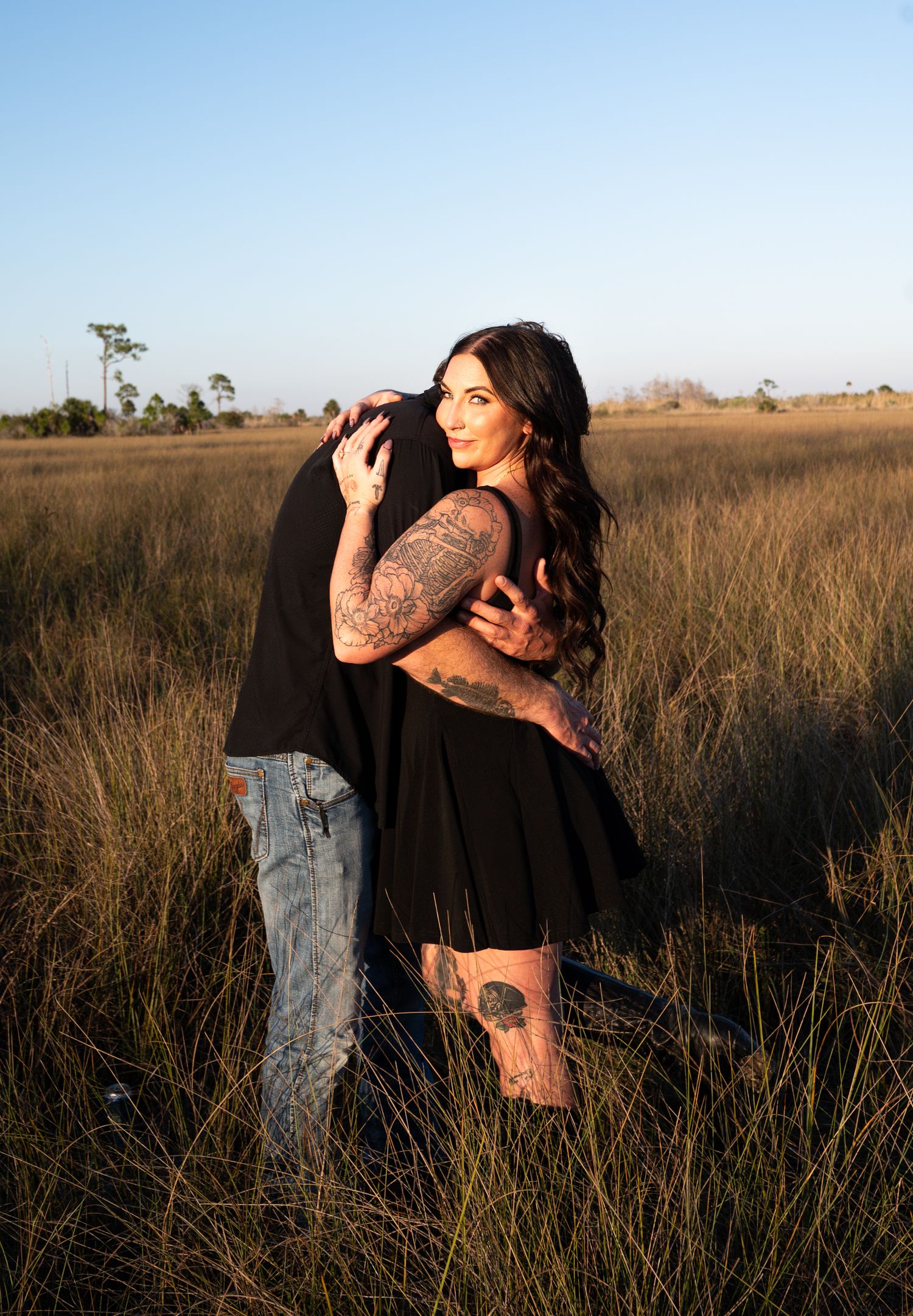 A couple embraces in a golden field during sunset as warm light bathes the tall grass and natural landscape.