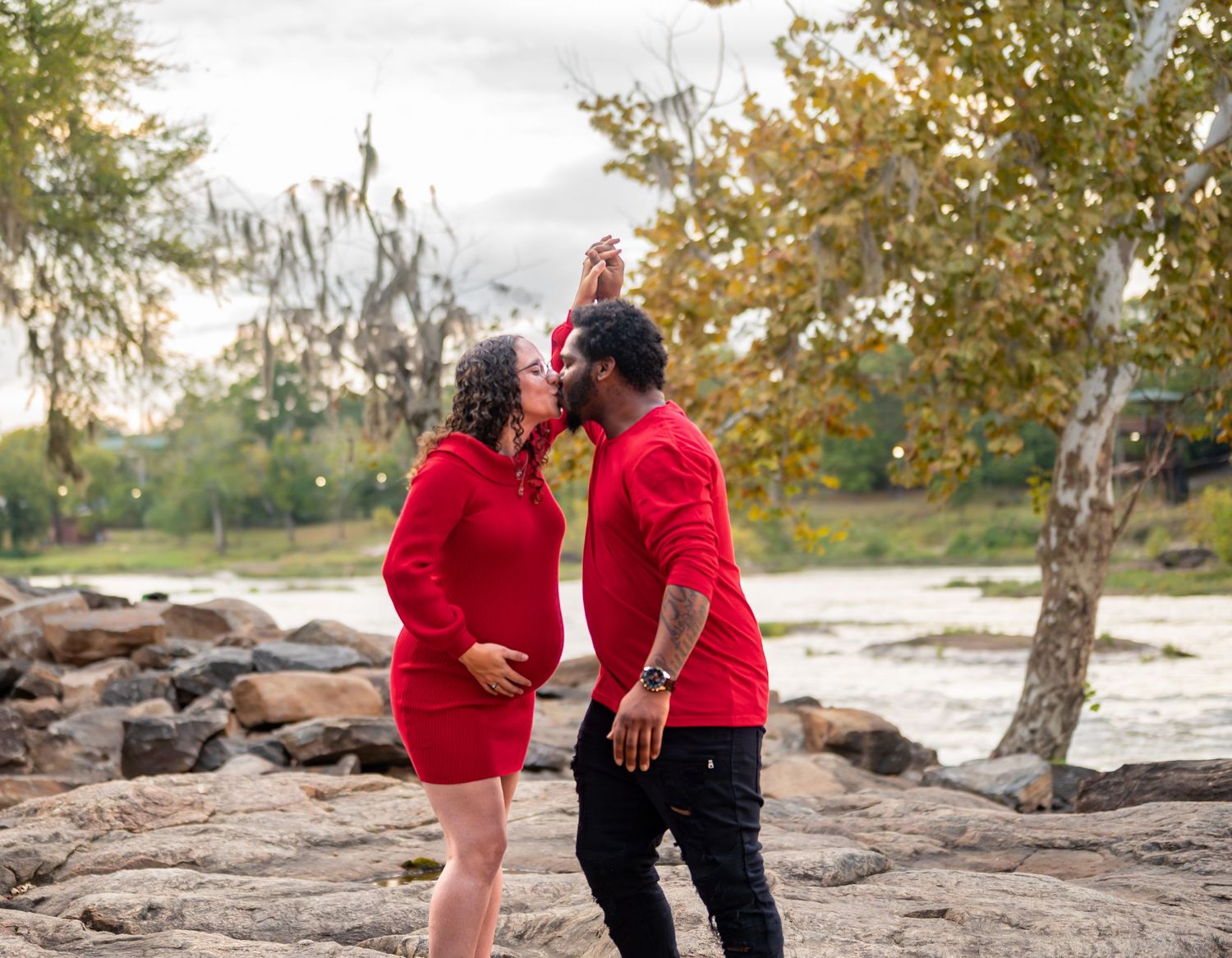 Couple in coordinating red outfits walks together along rocky waterfront during photo session.