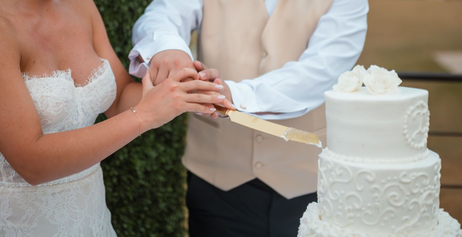 A white wedding cake with intricate lace details and decorative patterns sits on display at an outdoor reception.
