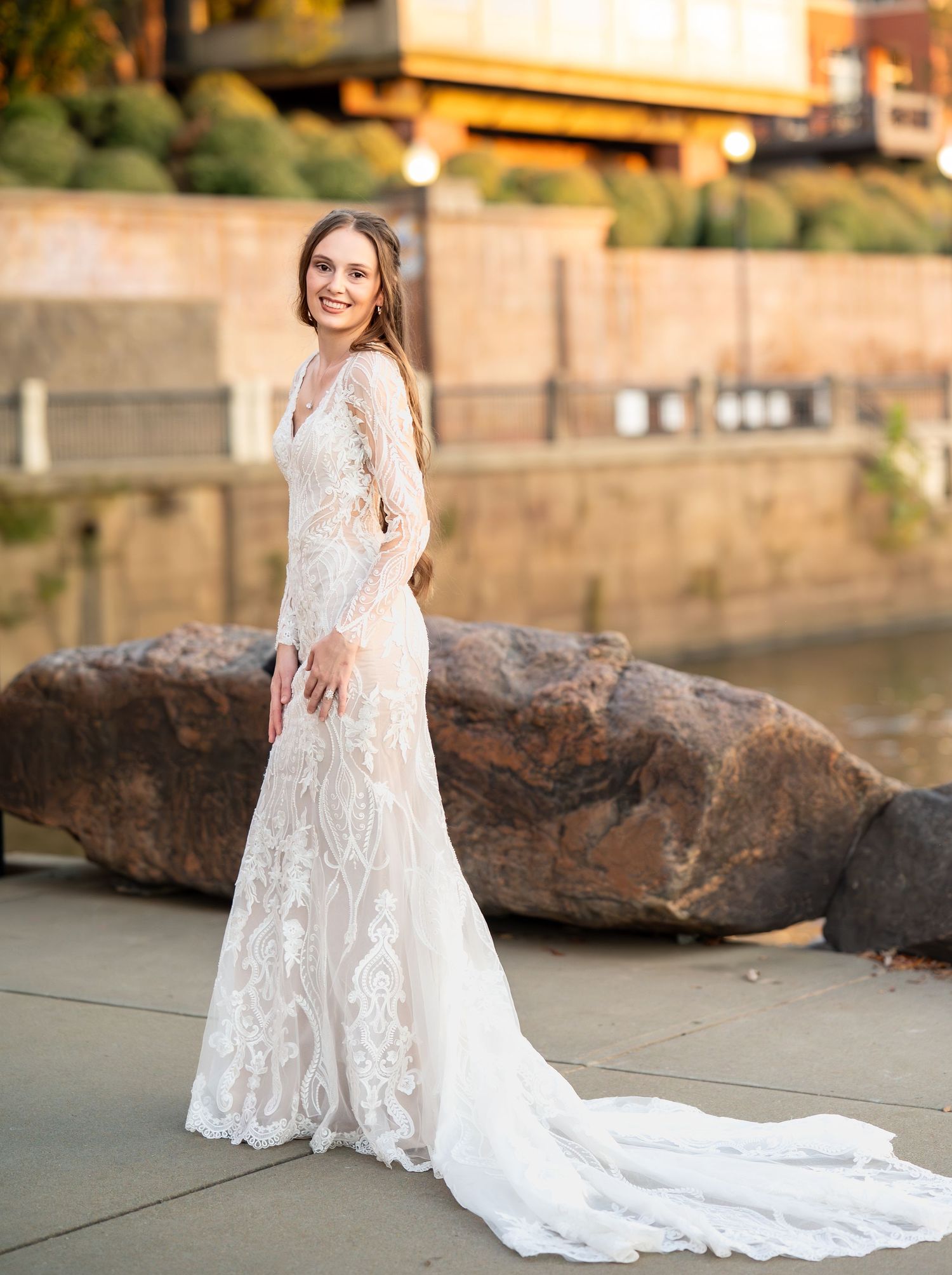 A beautiful lace wedding dress with a long flowing train is showcased along a waterfront setting.
