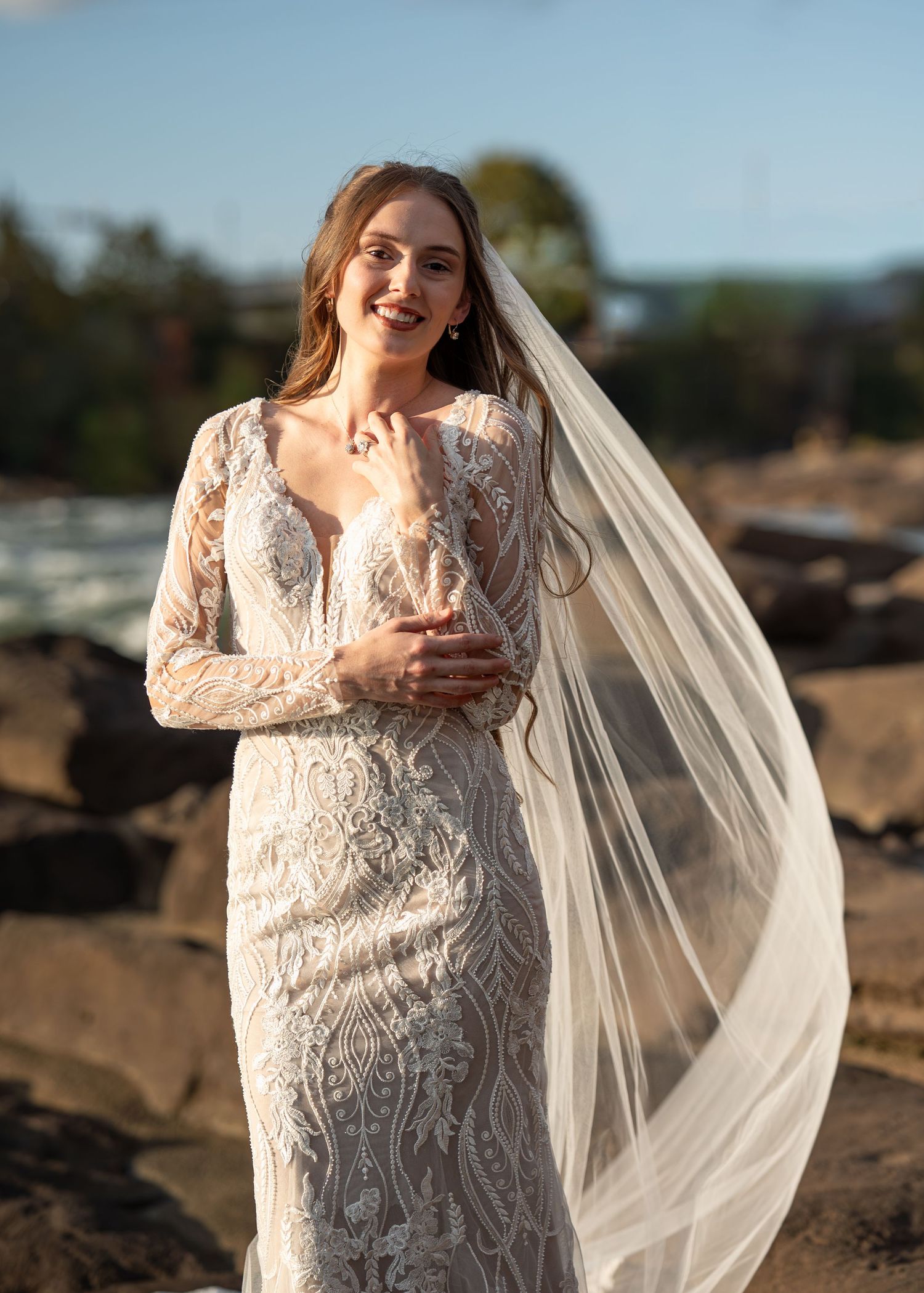 Elegant bridal portrait featuring a fitted lace gown with flowing veil against a natural outdoor backdrop.