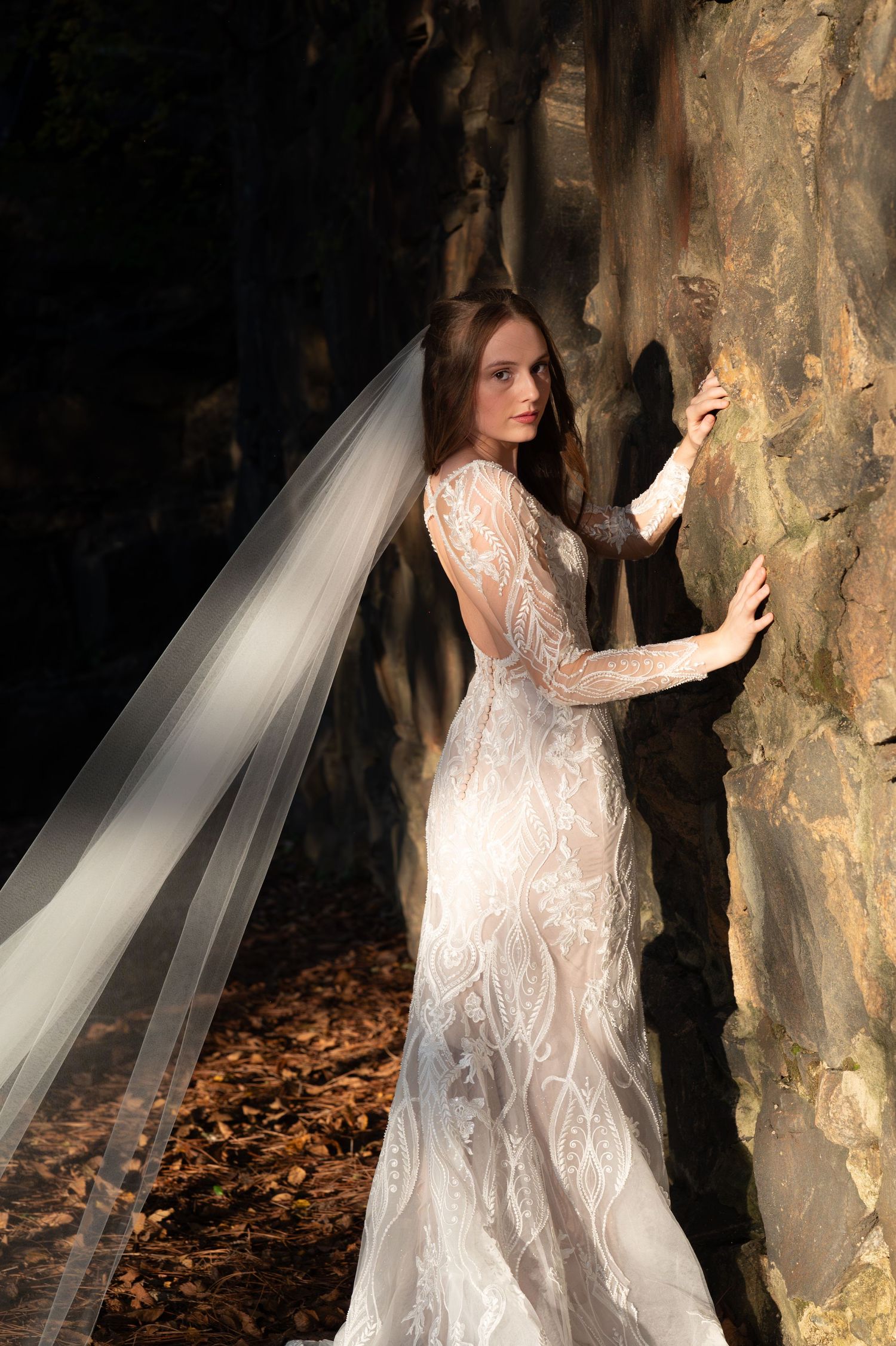 Ethereal bridal portrait showing a lace gown against textured stone wall with long flowing veil in natural light.