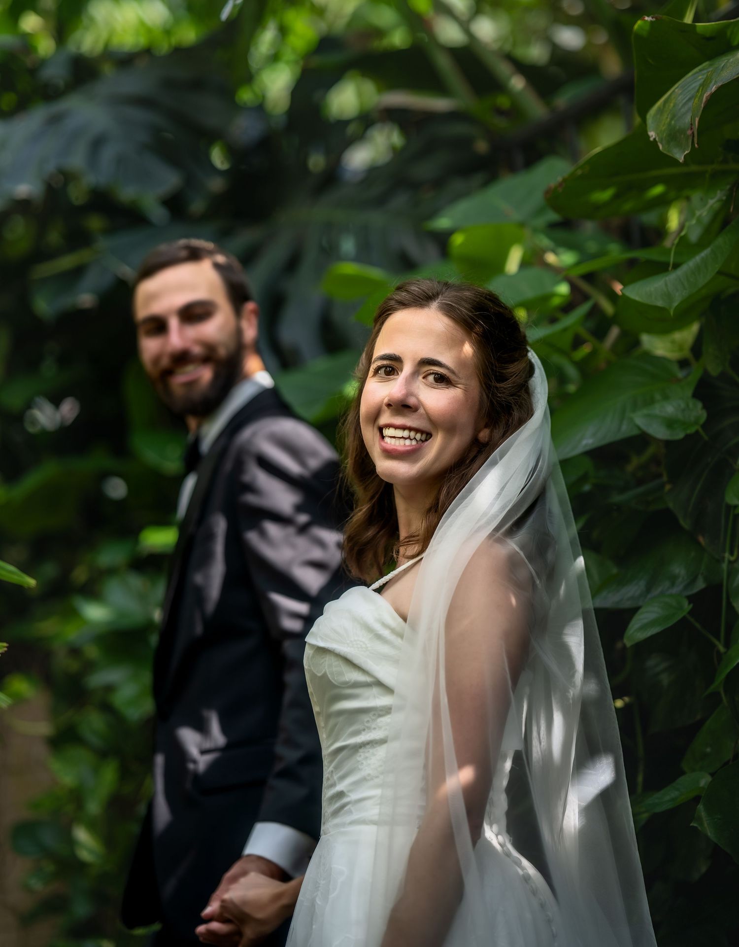 A joyful bride in a white dress and veil smiles while standing among lush green foliage with her groom in a dark suit.