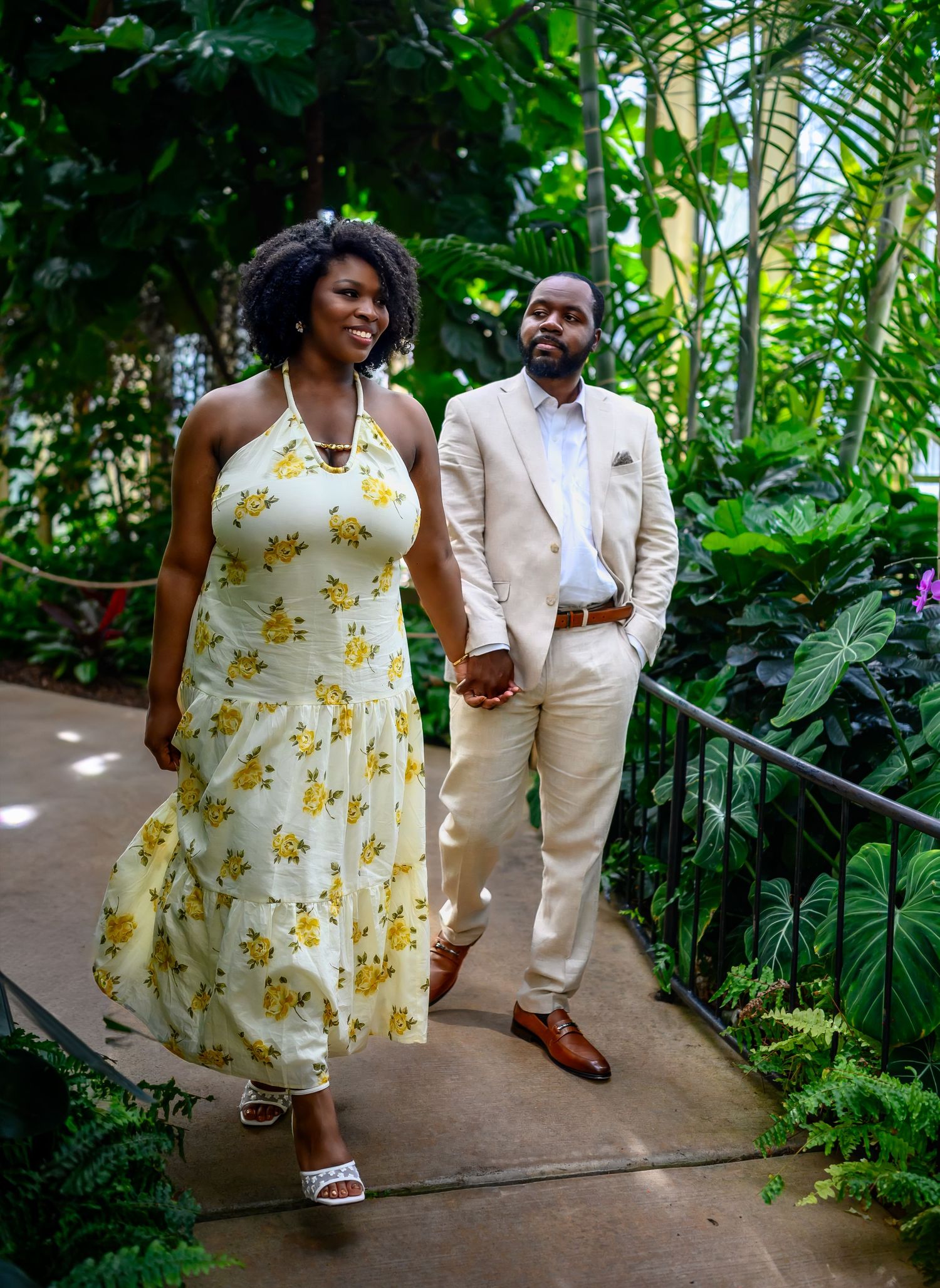 Engaged couple walking hand in hand through a lush tropical greenhouse garden pathway.