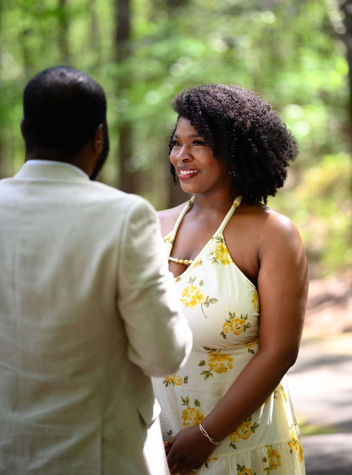 A smiling woman in a yellow floral halter dress stands outdoors in a wooded setting facing a man in a light suit.