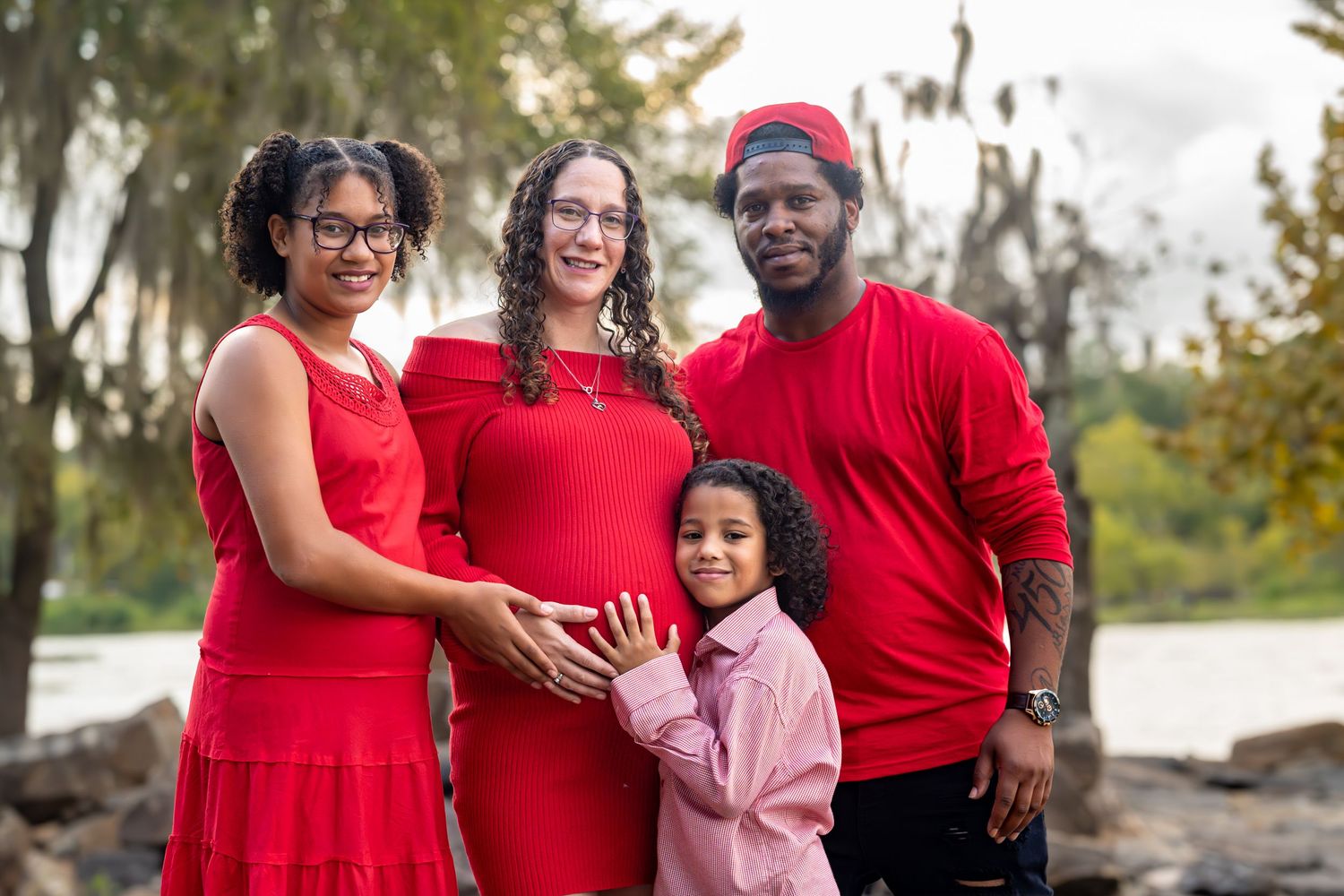 Happy family in red clothing poses together by the water during outdoor portrait session.