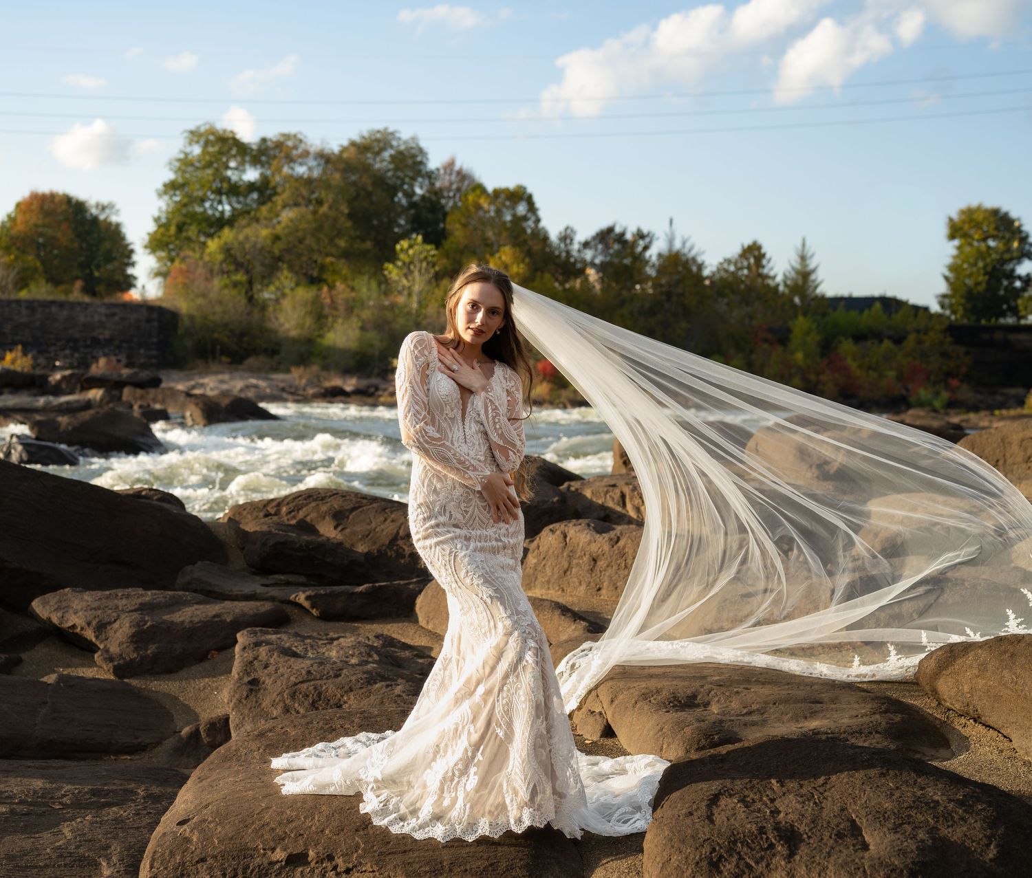 Wedding dress and veil dramatically captured in motion against rocky riverbank scenery.