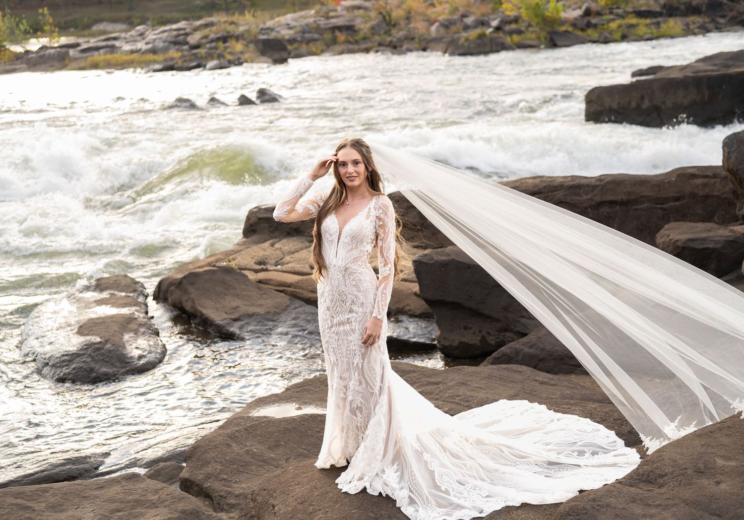 A bride in a flowing white dress stands on rocky coastal cliffs as her veil billows dramatically in the wind.