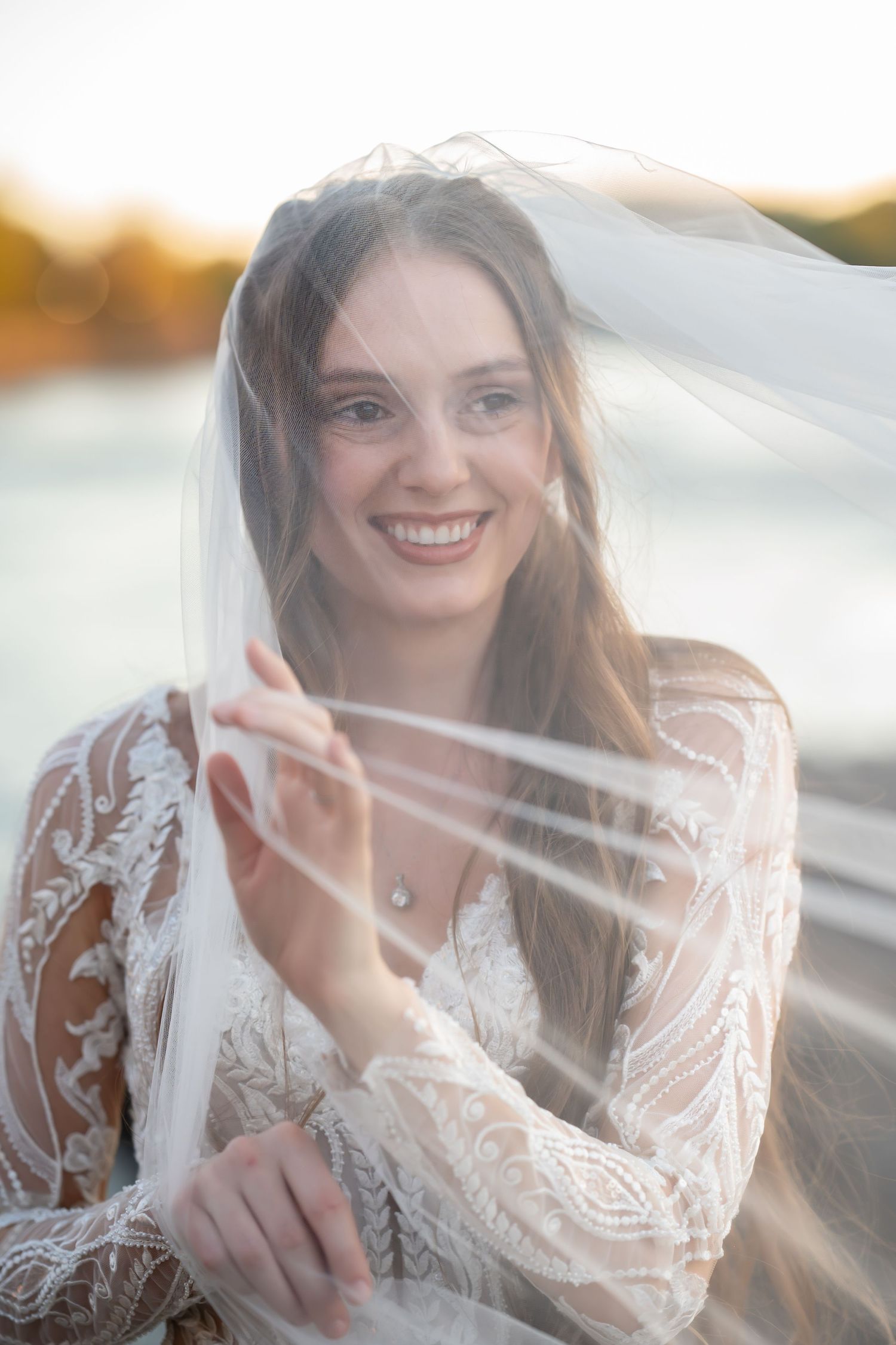 Joyful bride in lace gown plays with flowing veil during sunset photoshoot.