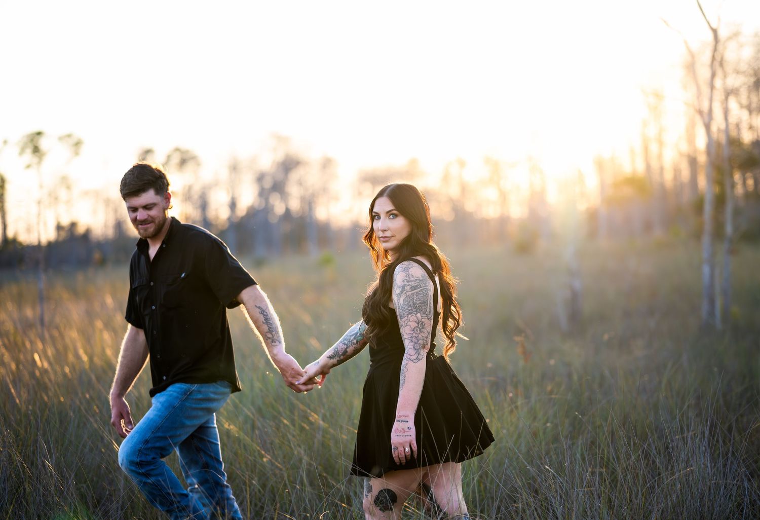 A couple walks hand in hand through a sunlit meadow at golden hour.