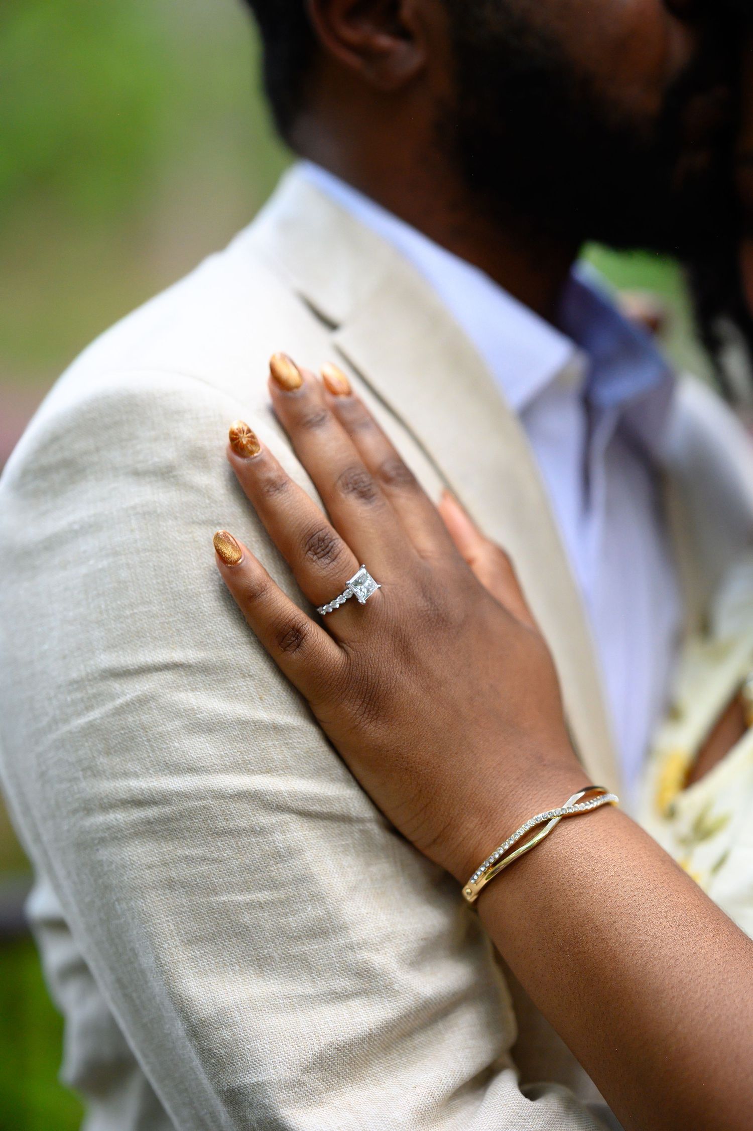 Close-up of brides hand showing diamond engagement ring with gold nails resting on grooms beige suit.