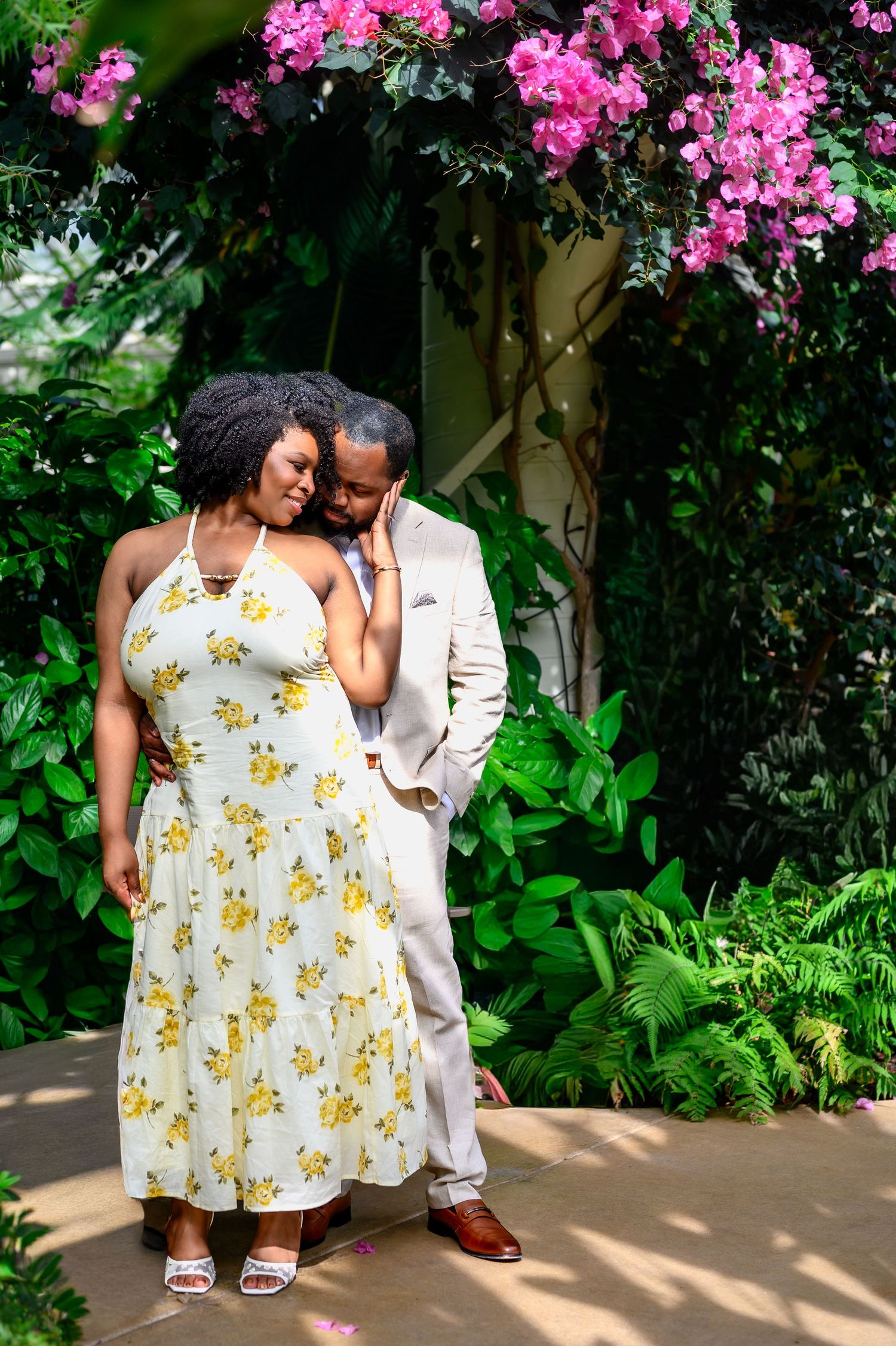 A romantic couple stands close together beneath vibrant pink bougainvillea blooms in a lush garden.