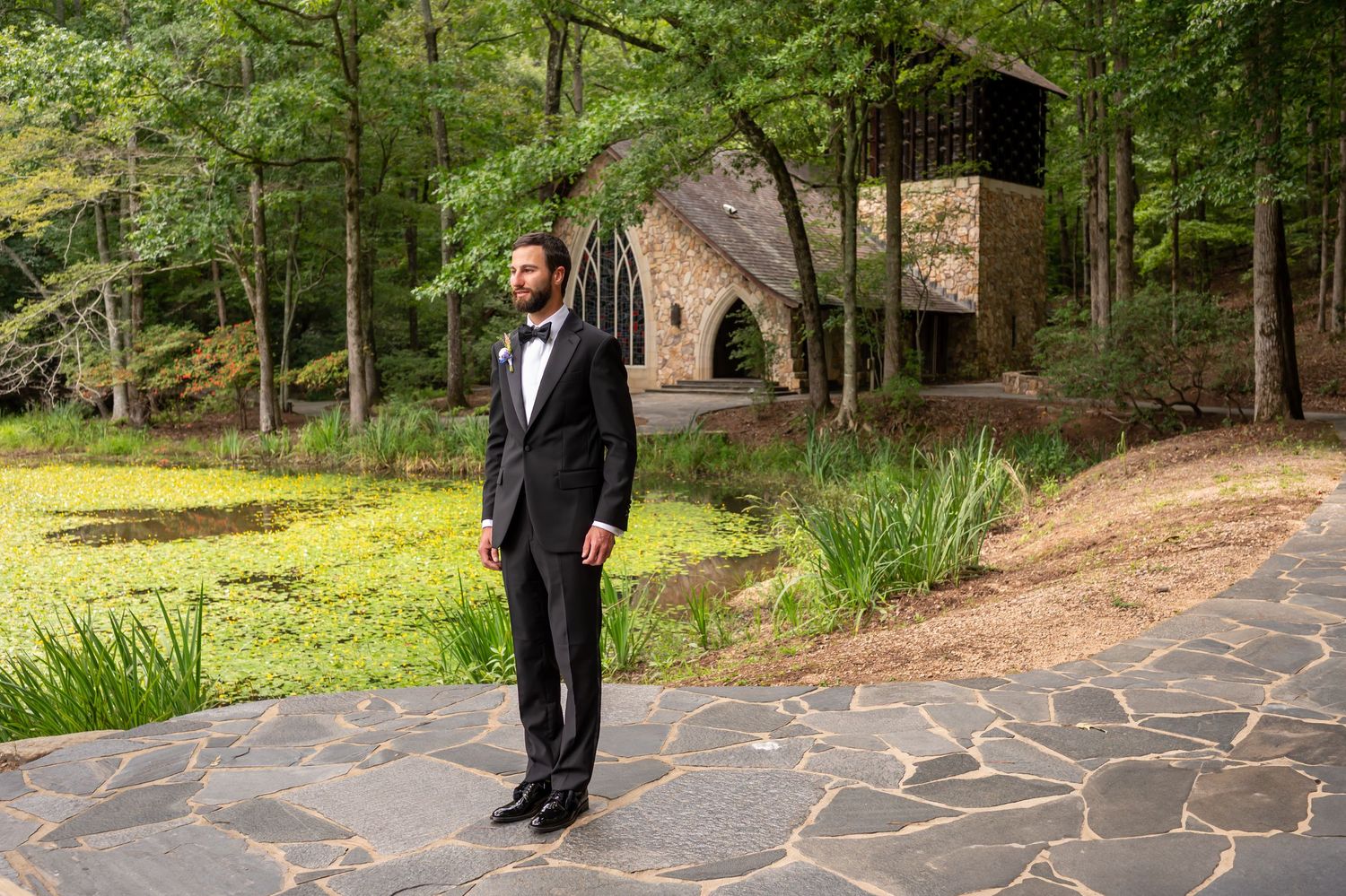 A person in a formal black tuxedo stands on a stone path near a rustic chapel surrounded by lush green forest.