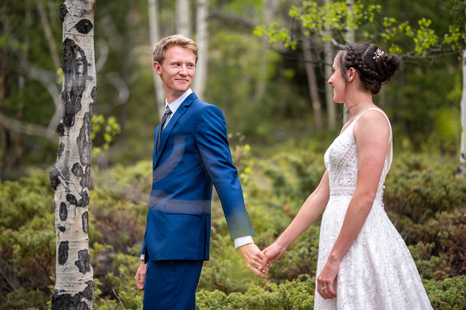 A couple holding hands in a forest setting with aspen trees and greenery surrounding them.