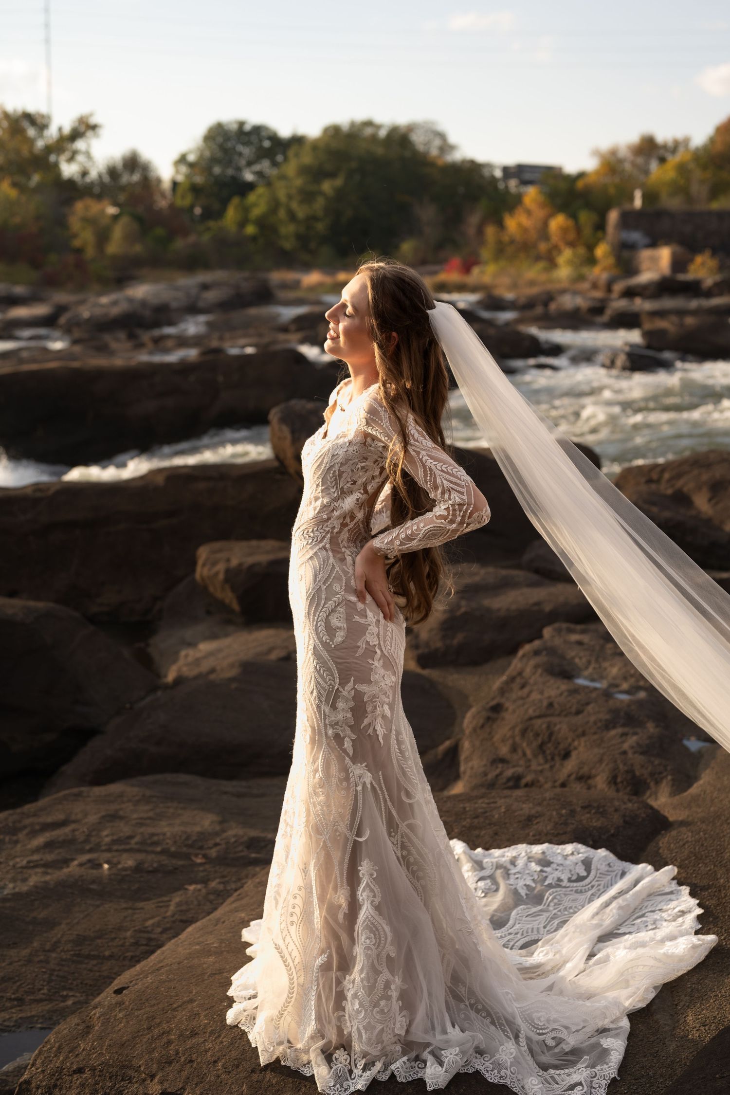 Elegant wedding dress flowing across rocky terrain near a river with autumn foliage in the background.