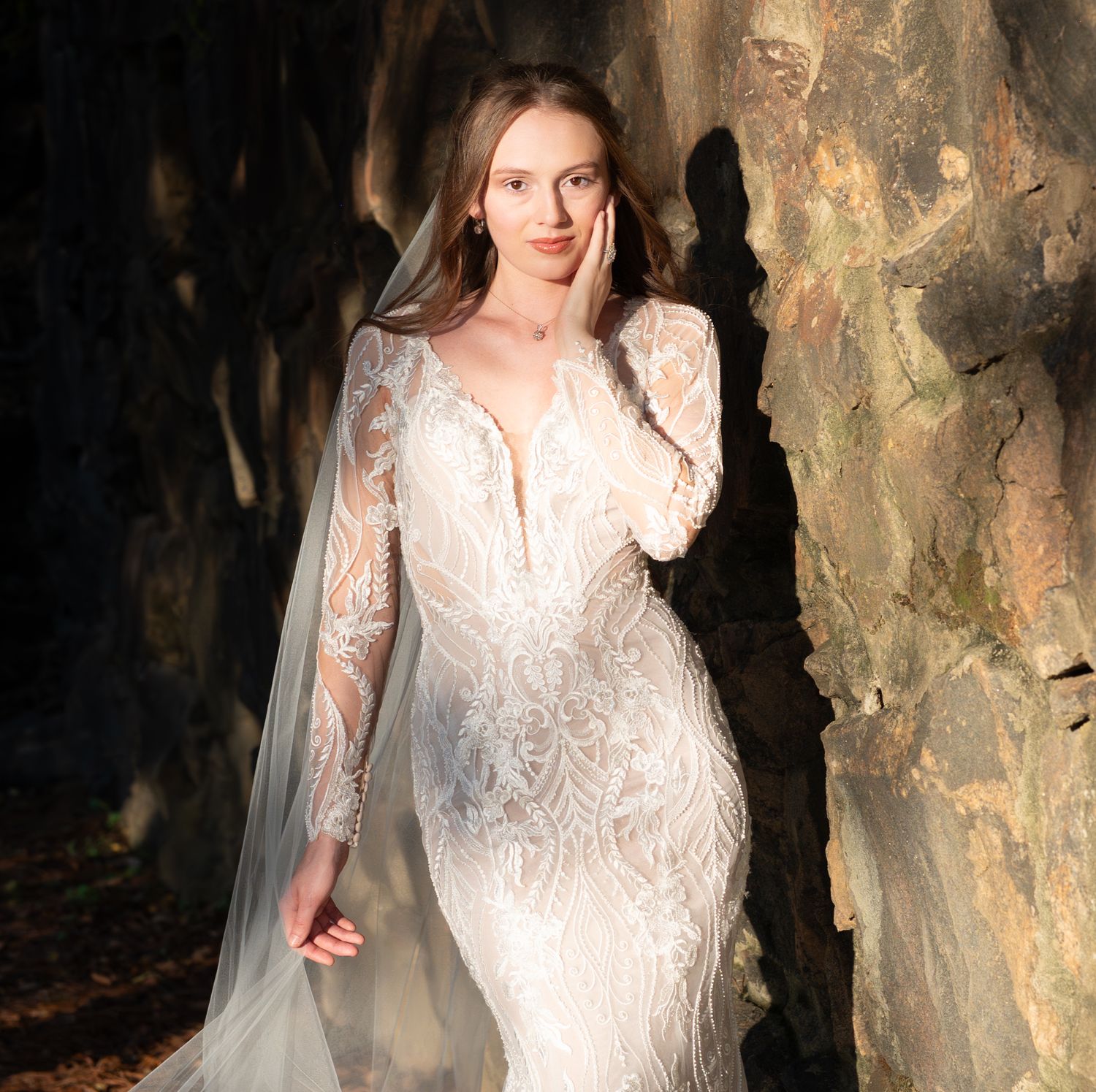 A bride poses in an intricate lace gown against a textured stone wall backdrop.