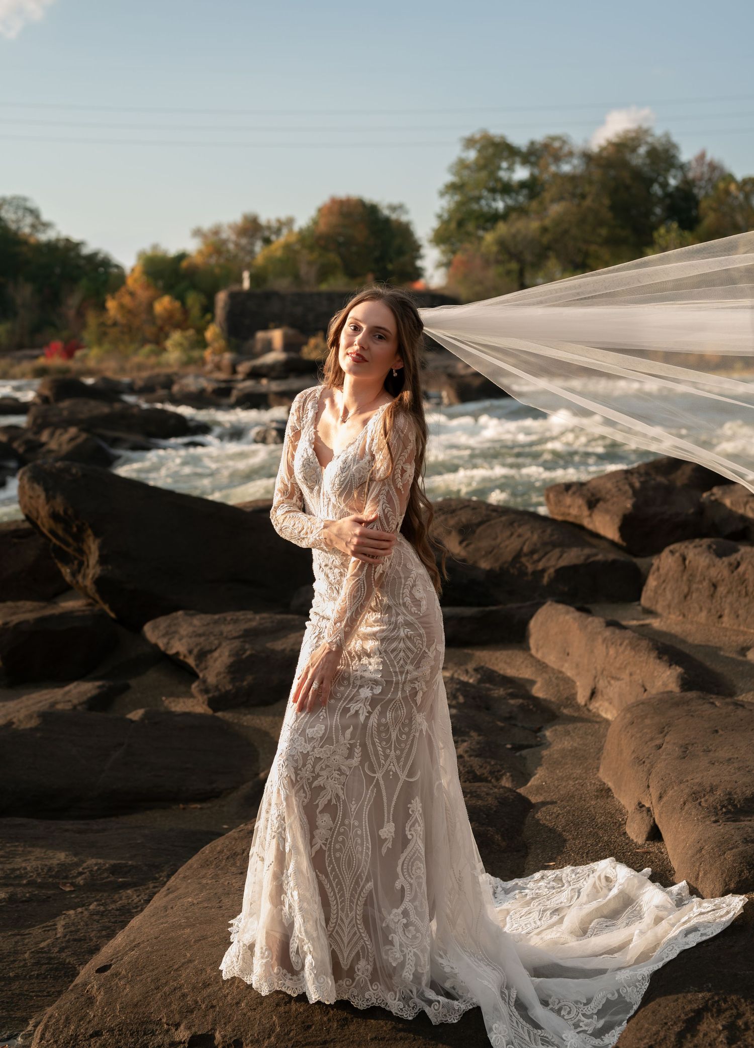 Ethereal bridal photography session on rocky waterfront with flowing veil and lace gown.