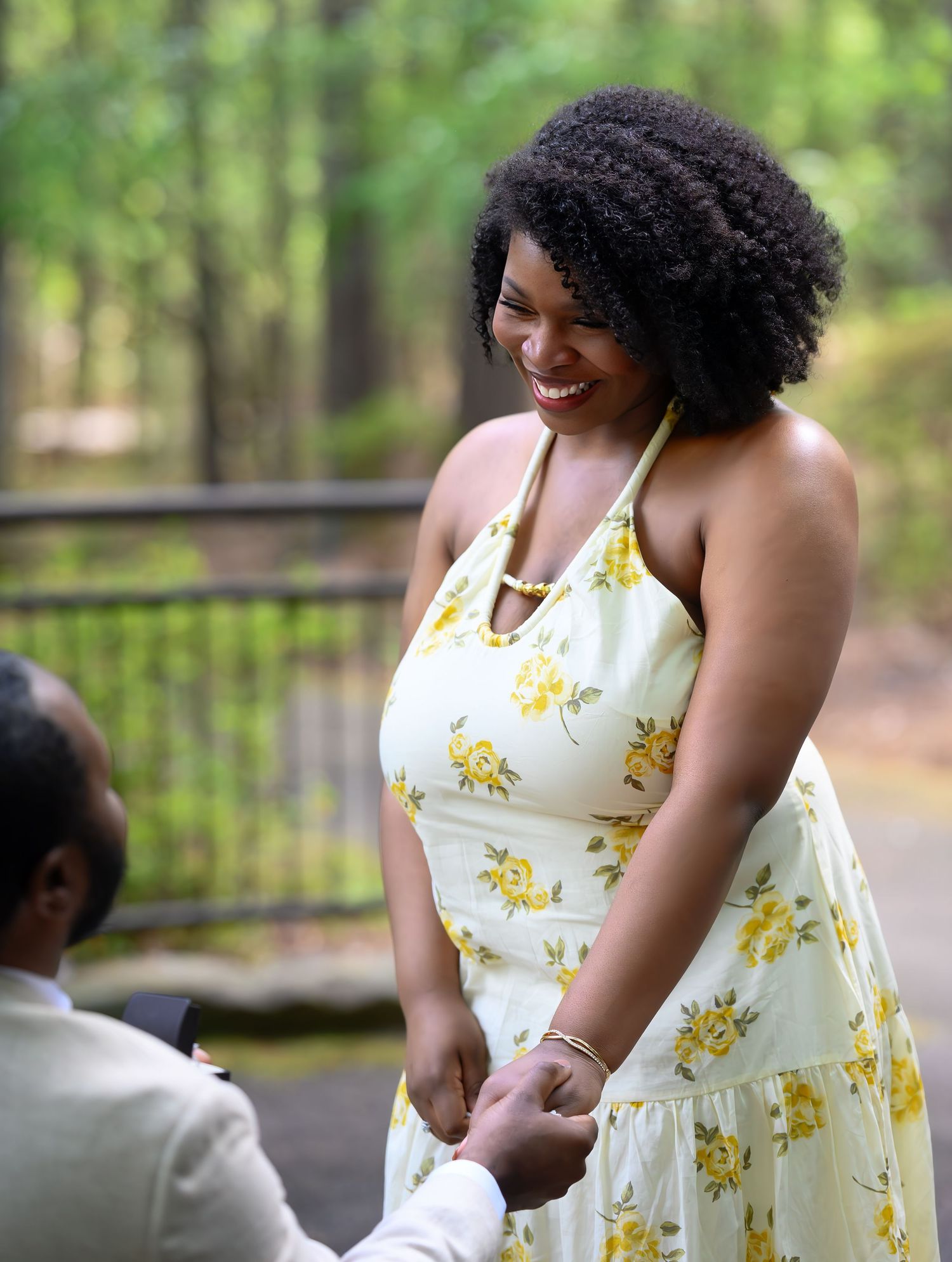 Joyful woman in a yellow floral dress smiling as her partner holds her hands during a proposal.