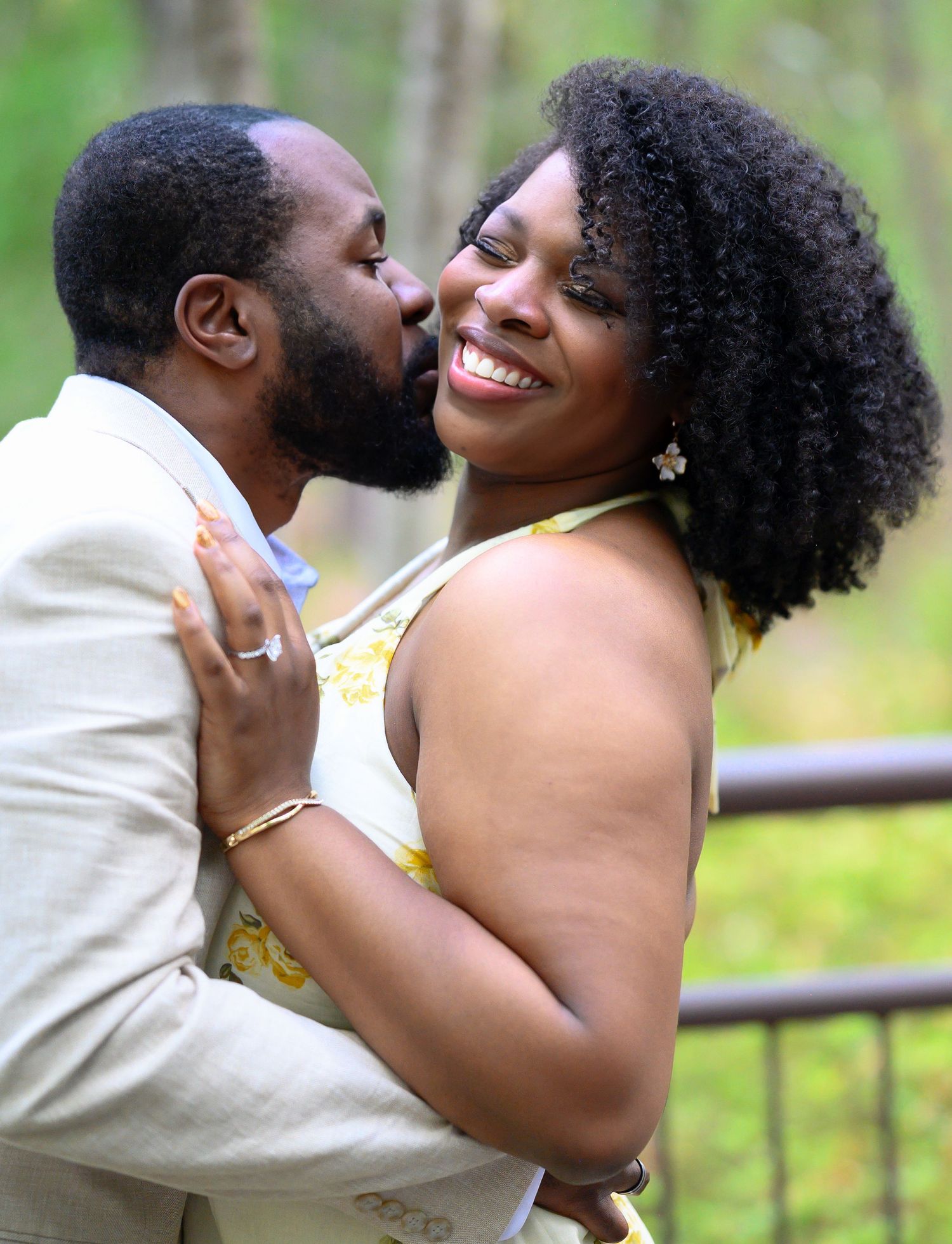 A man kisses a smiling womans cheek as she laughs joyfully outdoors with a soft green wooded background.