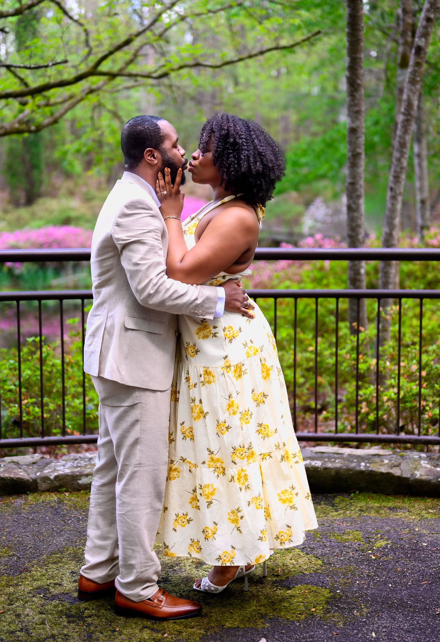 A romantic couple sharing a kiss outdoors in a garden with colorful azaleas blooming behind them.