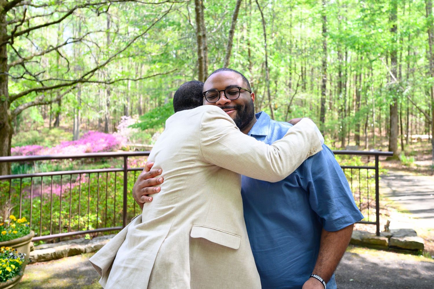 Two men sharing a warm embrace outdoors in a blooming garden with azaleas in the background.