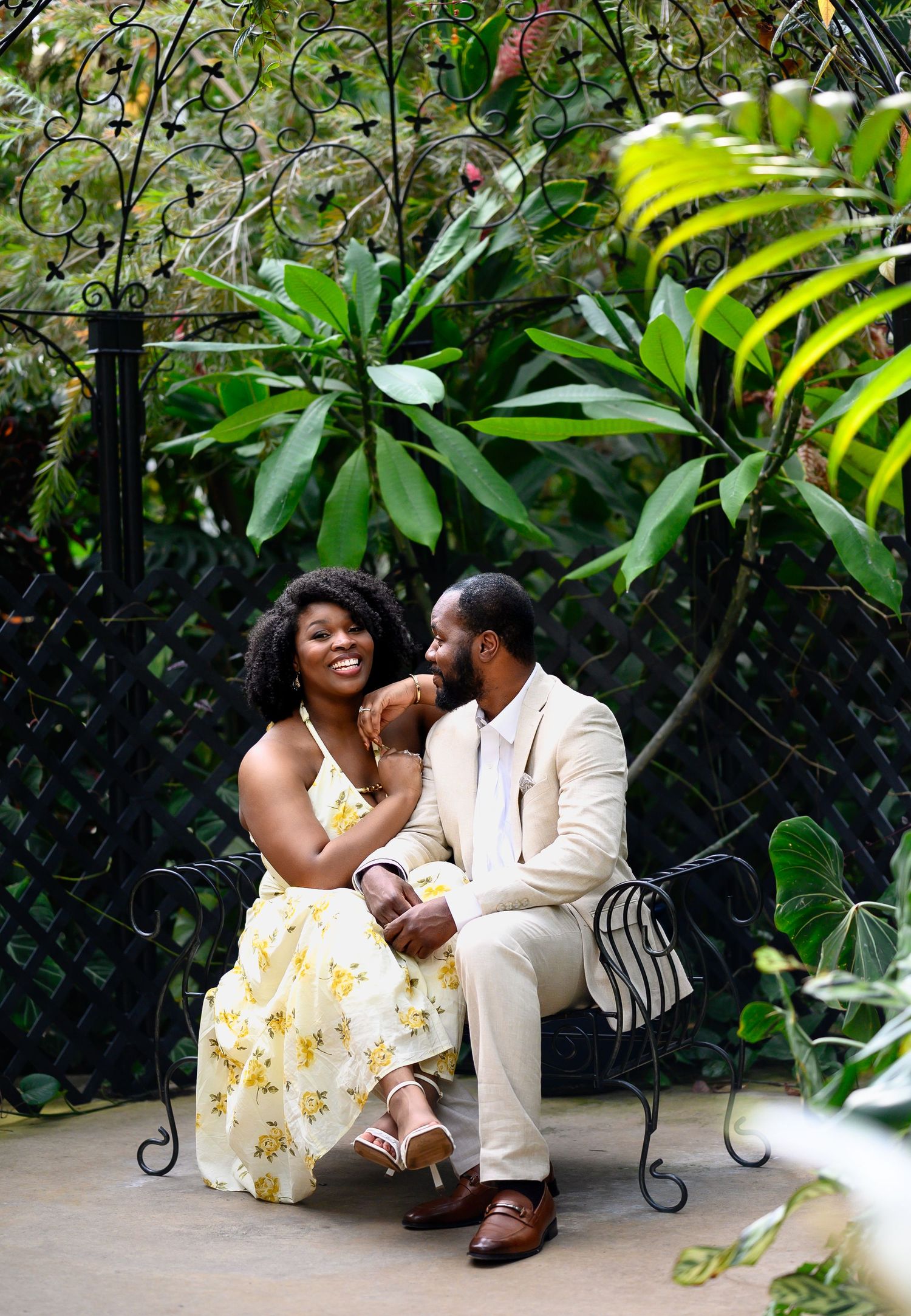 A happy couple sits together on an ornate iron bench surrounded by tropical greenery, smiling warmly.