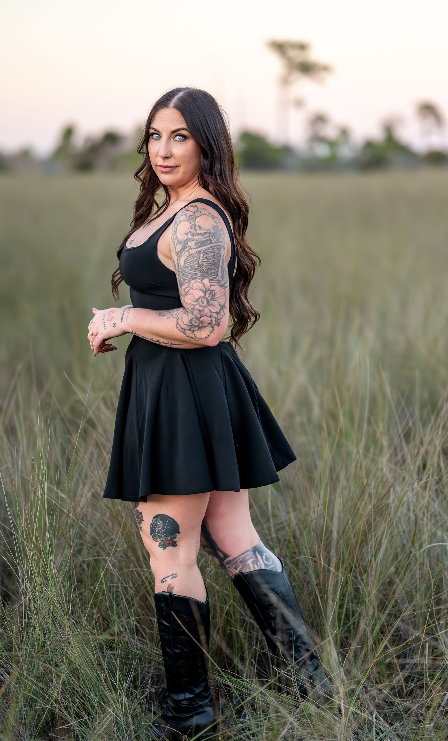A woman in a black dress and boots poses in a grassy field at sunset, showing off her artistic tattoos.