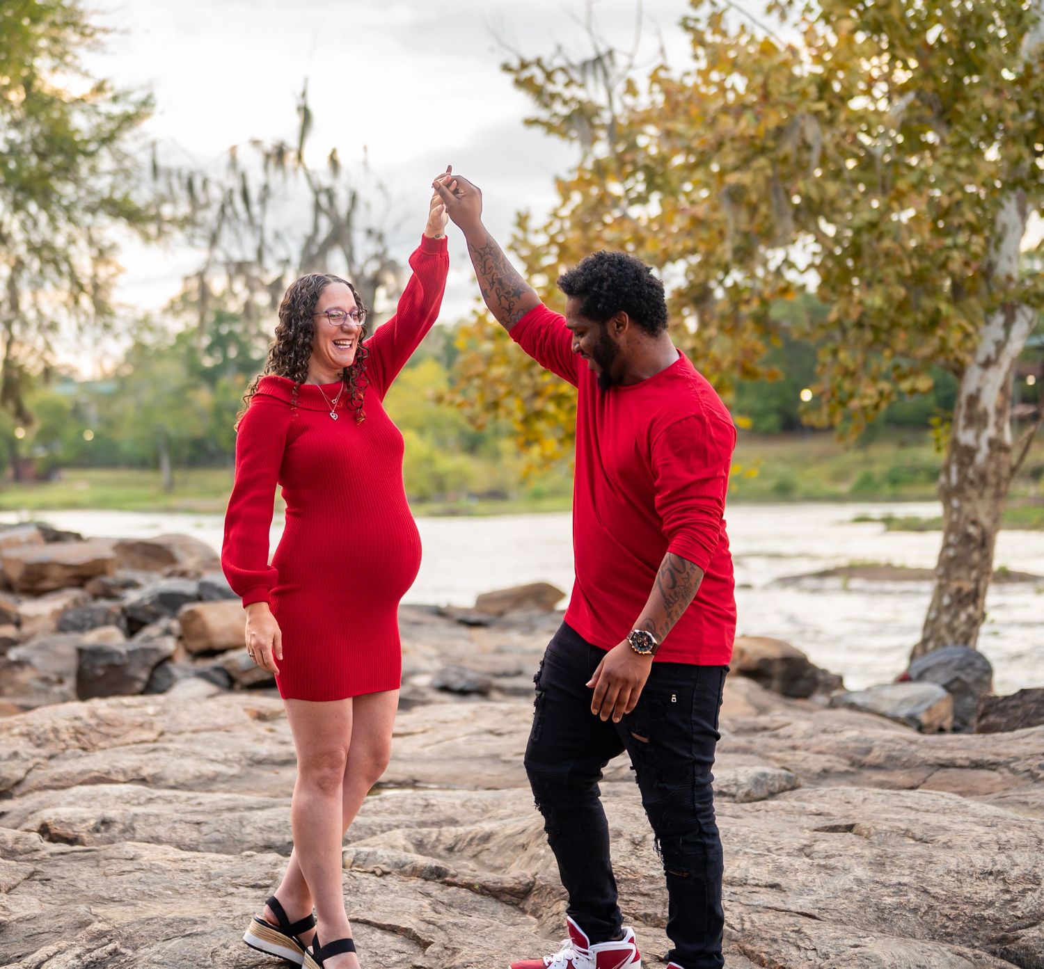 Family poses on rocky riverbank in red outfits during autumn maternity session.