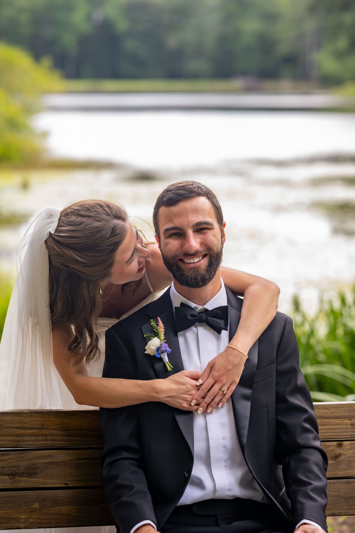 Newlyweds share intimate moment on wooden bench by lakeside.