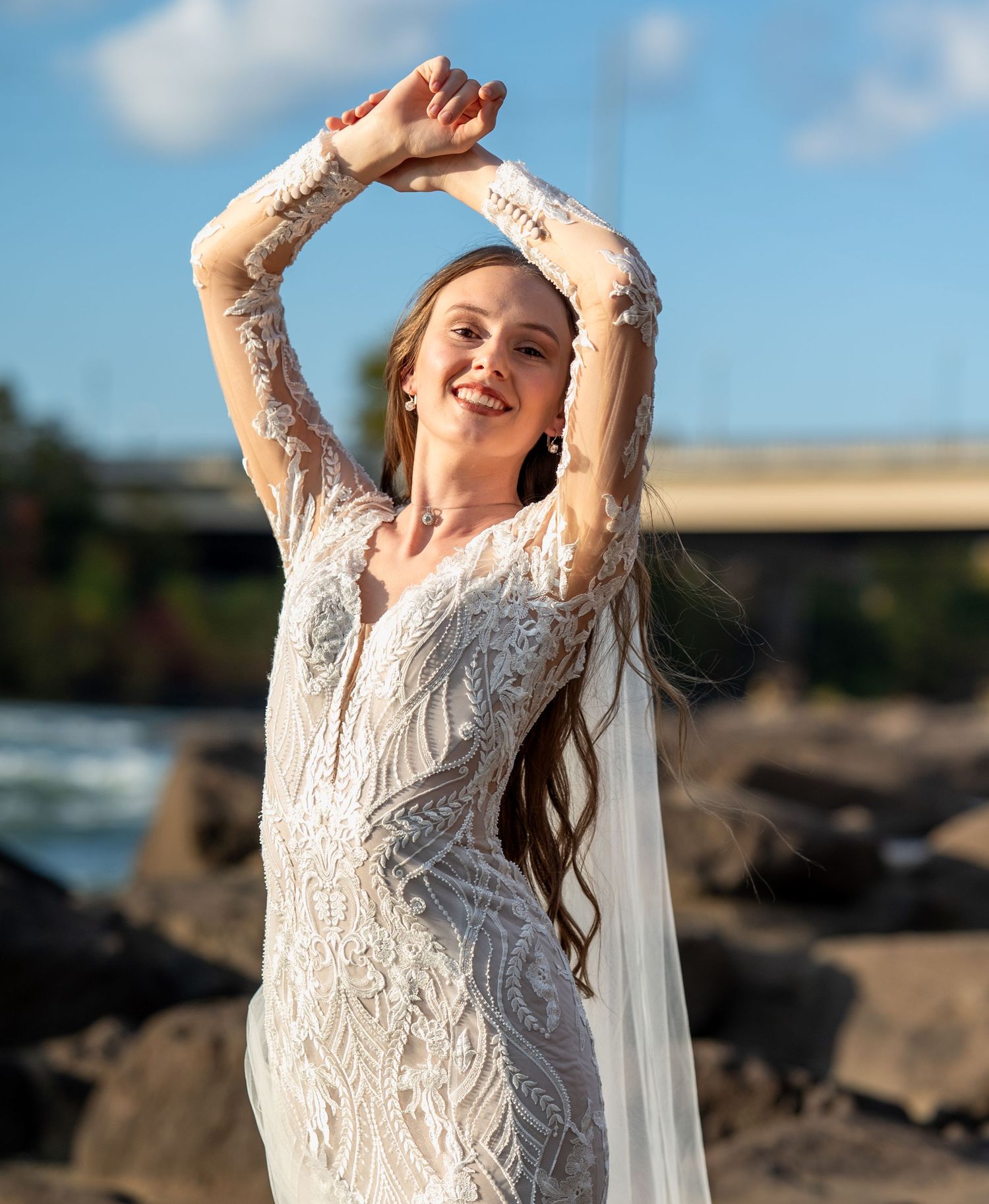 A woman in a flowing white lace dress dances joyfully on a rocky beach against a bright blue sky.