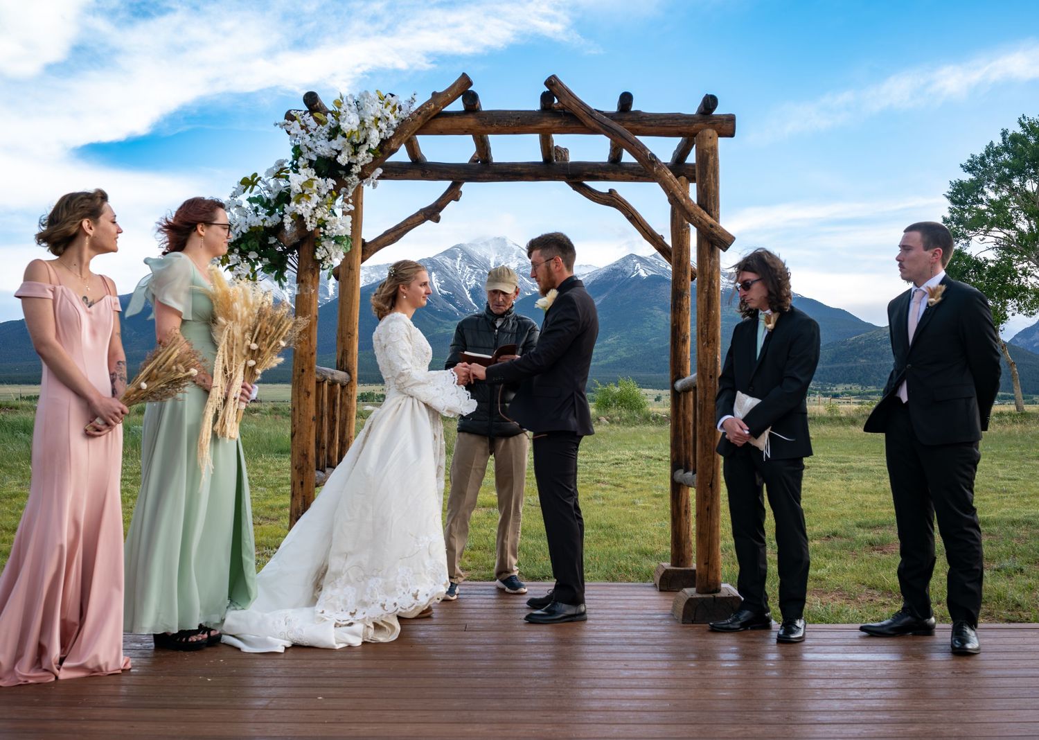 Wedding party gathered on wooden platform for ceremony with mountains and blue sky in background at the wedding venue, Barn at Sunset Ranch.