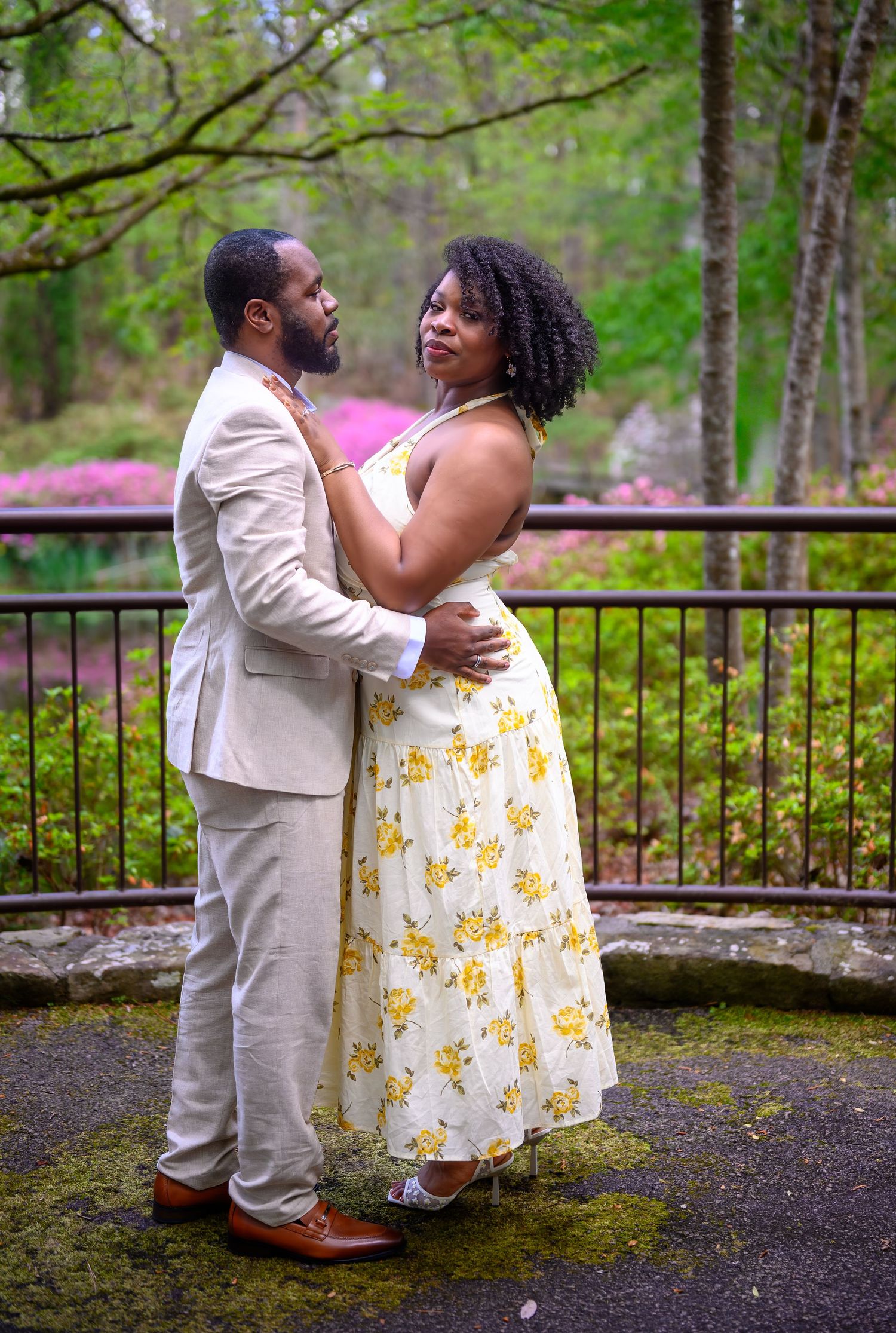 Couple embracing romantically in lush garden with blooming azaleas, man in beige suit and woman in yellow floral dress.
