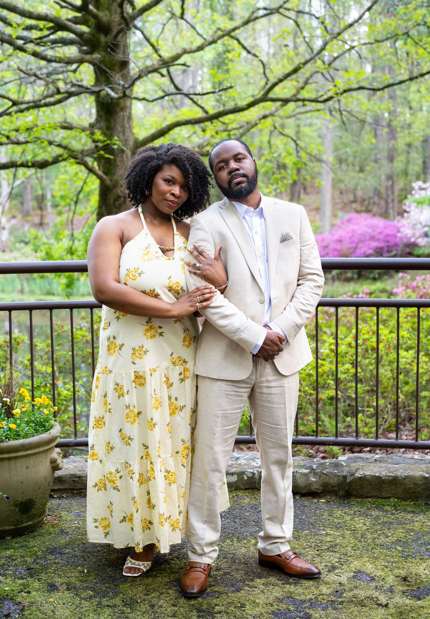 Elegant couple poses together on a stone patio, man in beige suit and woman in yellow floral halter dress.