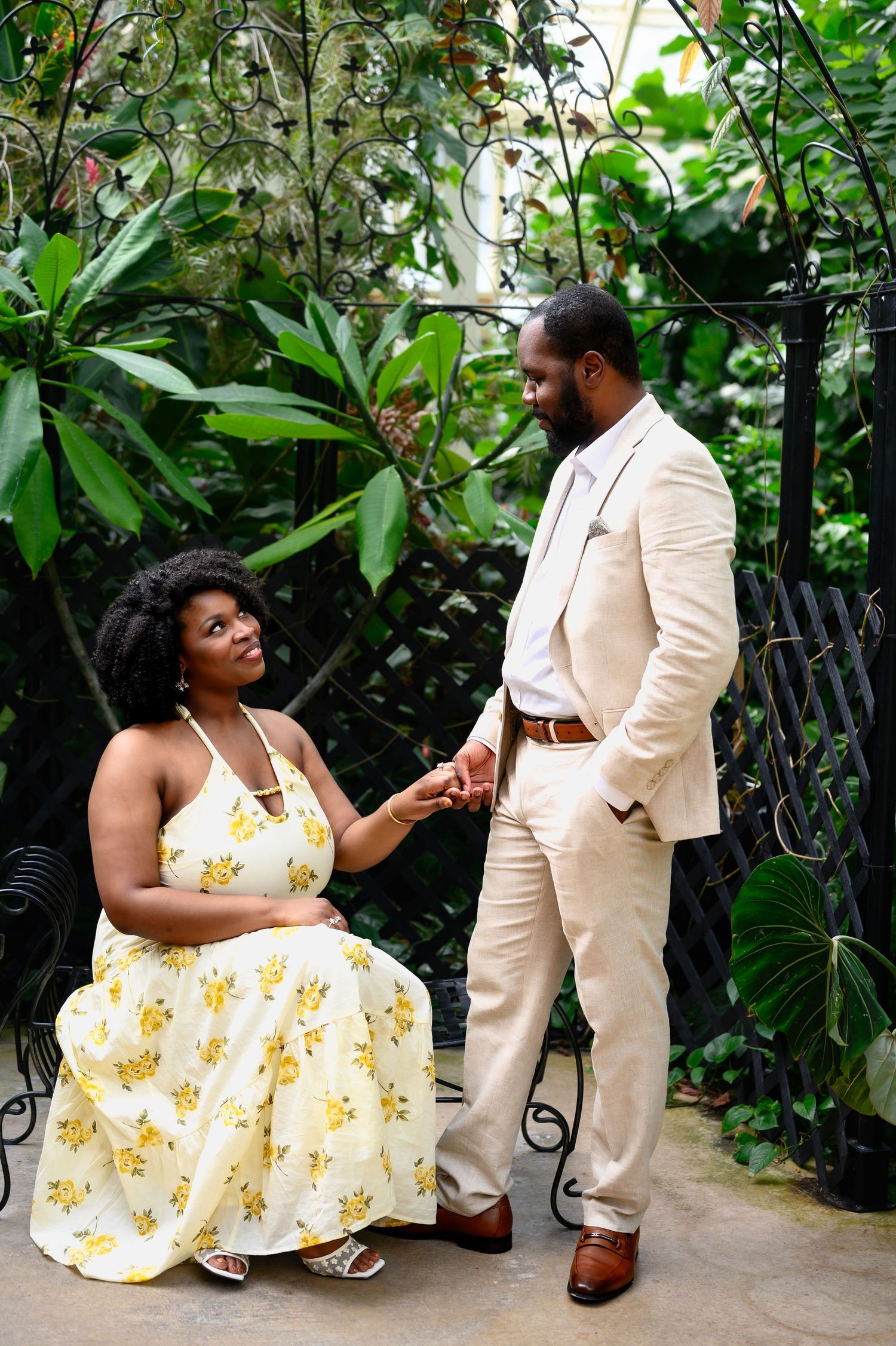 A woman in a yellow floral dress sits and holds hands with a man in a beige suit in a lush botanical garden.