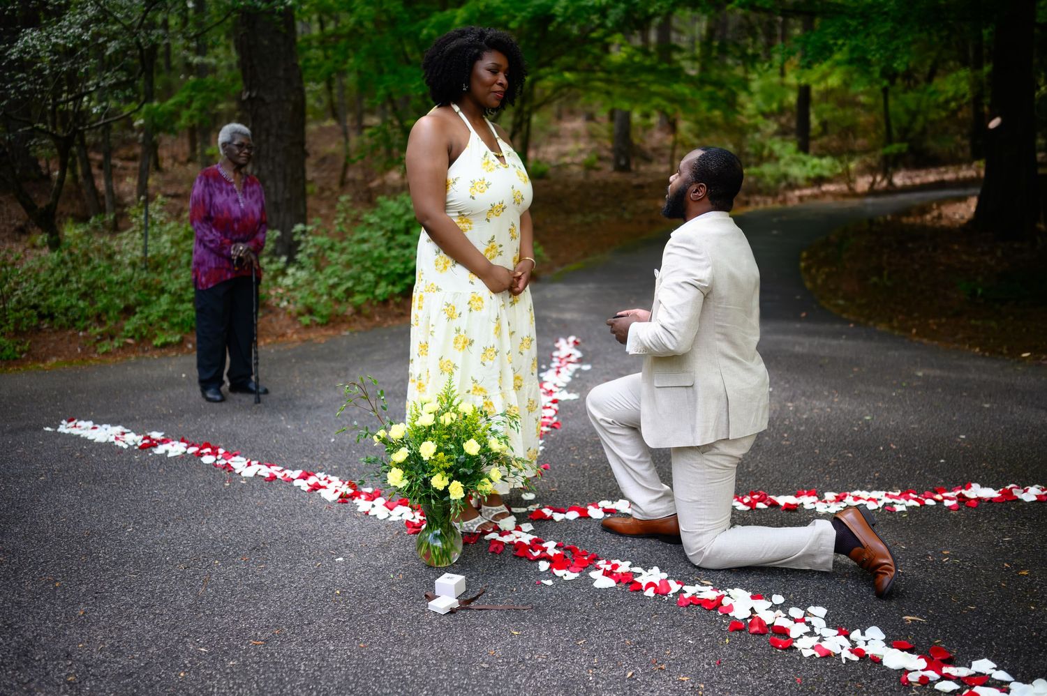 Man in white suit kneeling on one knee proposing to woman in yellow dress on rose petal path in forest.