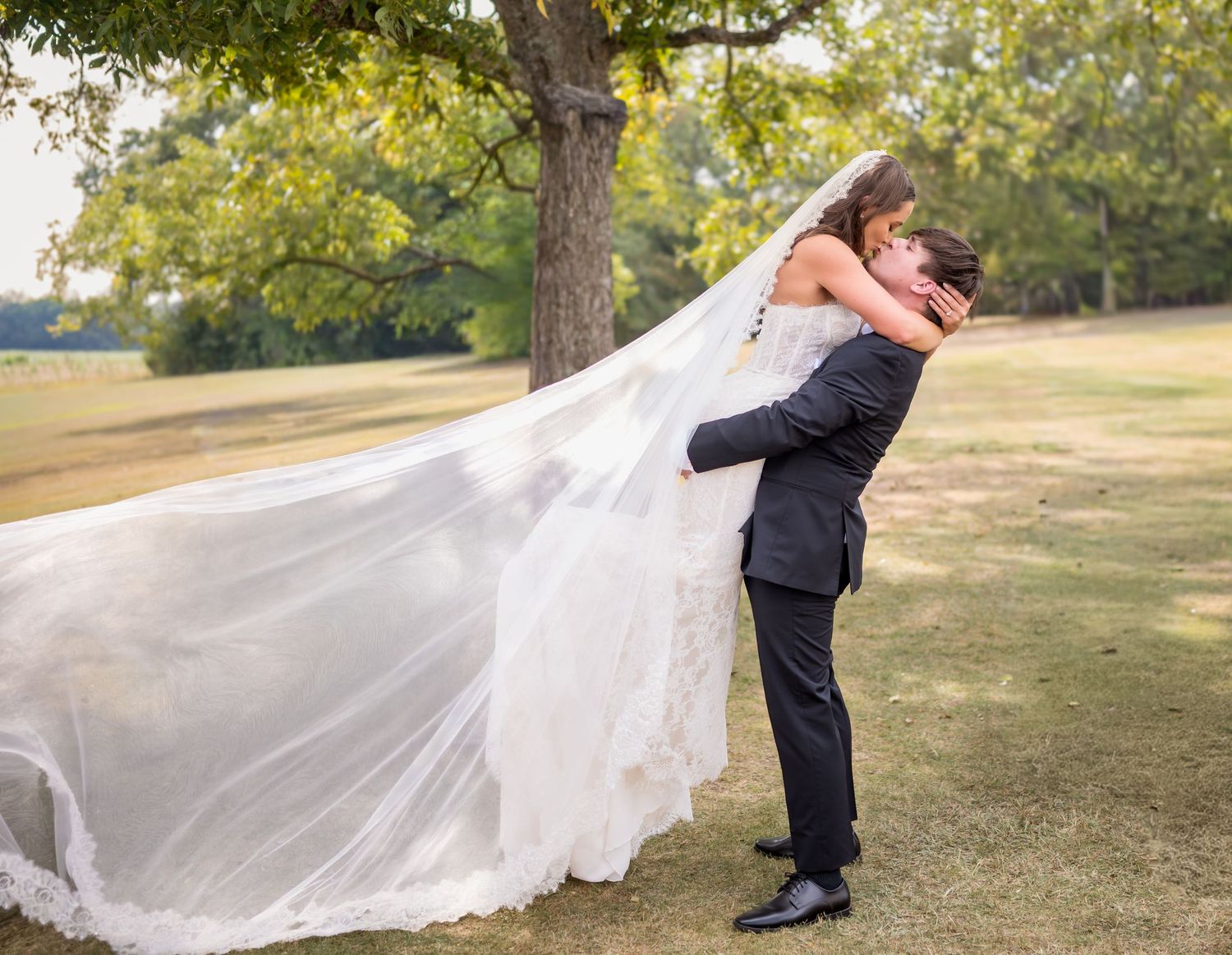 A romantic couple in wedding attire embraces in a grassy field while a flowing veil catches the breeze.