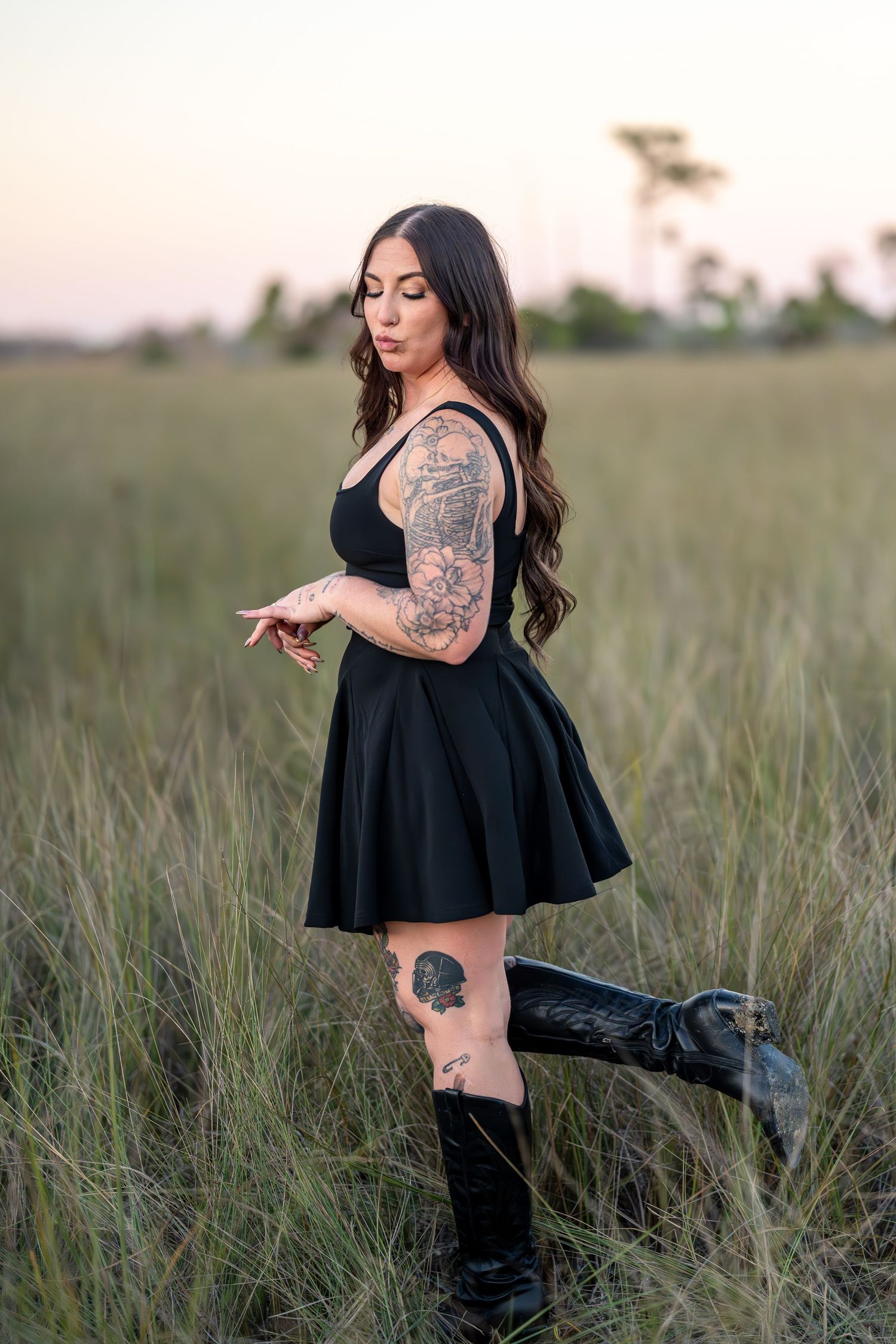 A person in a black dress and boots poses in a field of tall grass during sunset, showing off artistic tattoos.