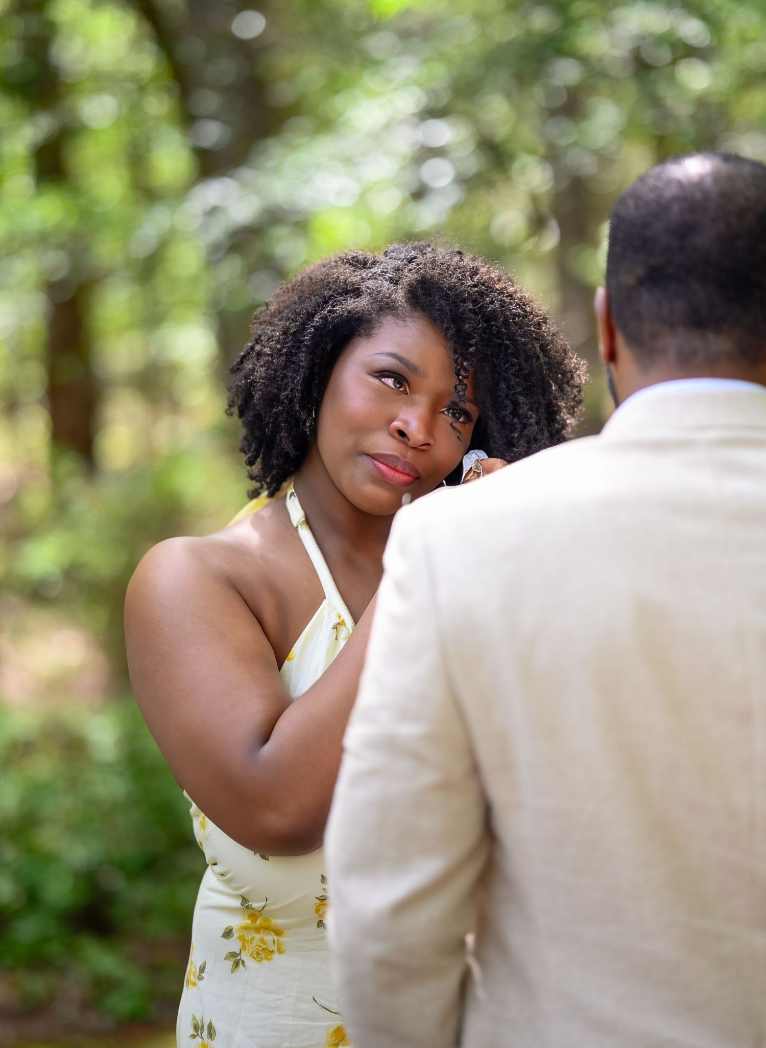 A woman in a yellow floral dress gazes lovingly at a man in a beige suit in a lush green forest.