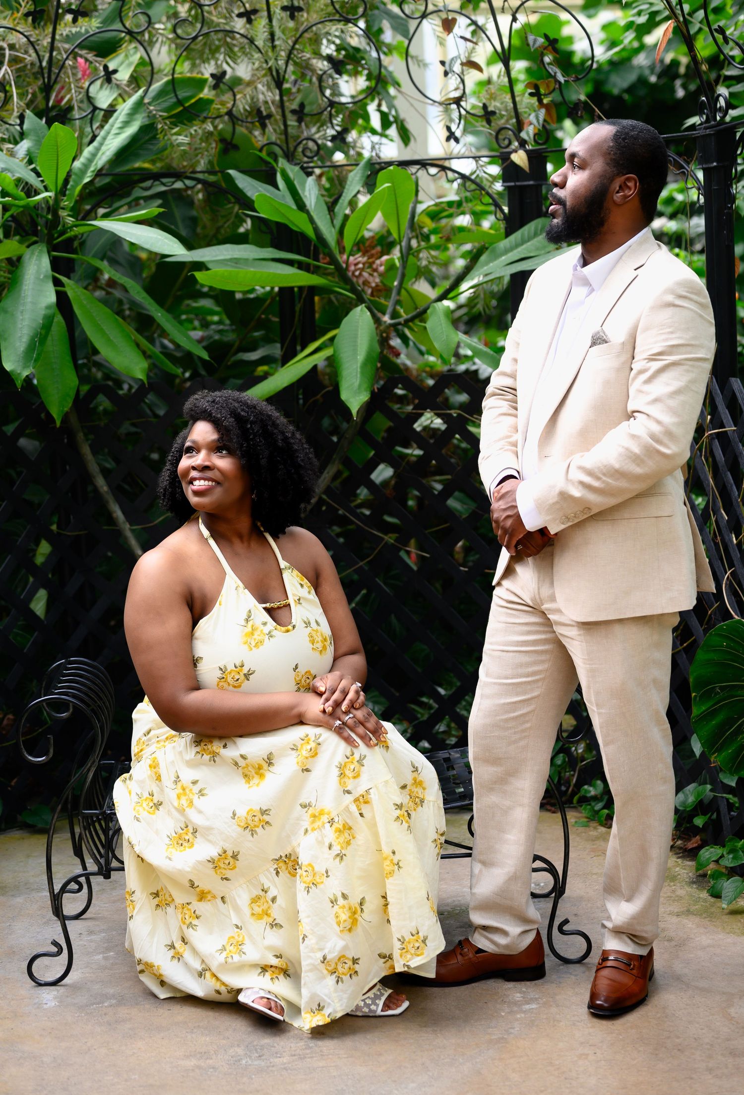 Woman in floral dress seated beside man in beige suit among tropical garden greenery.