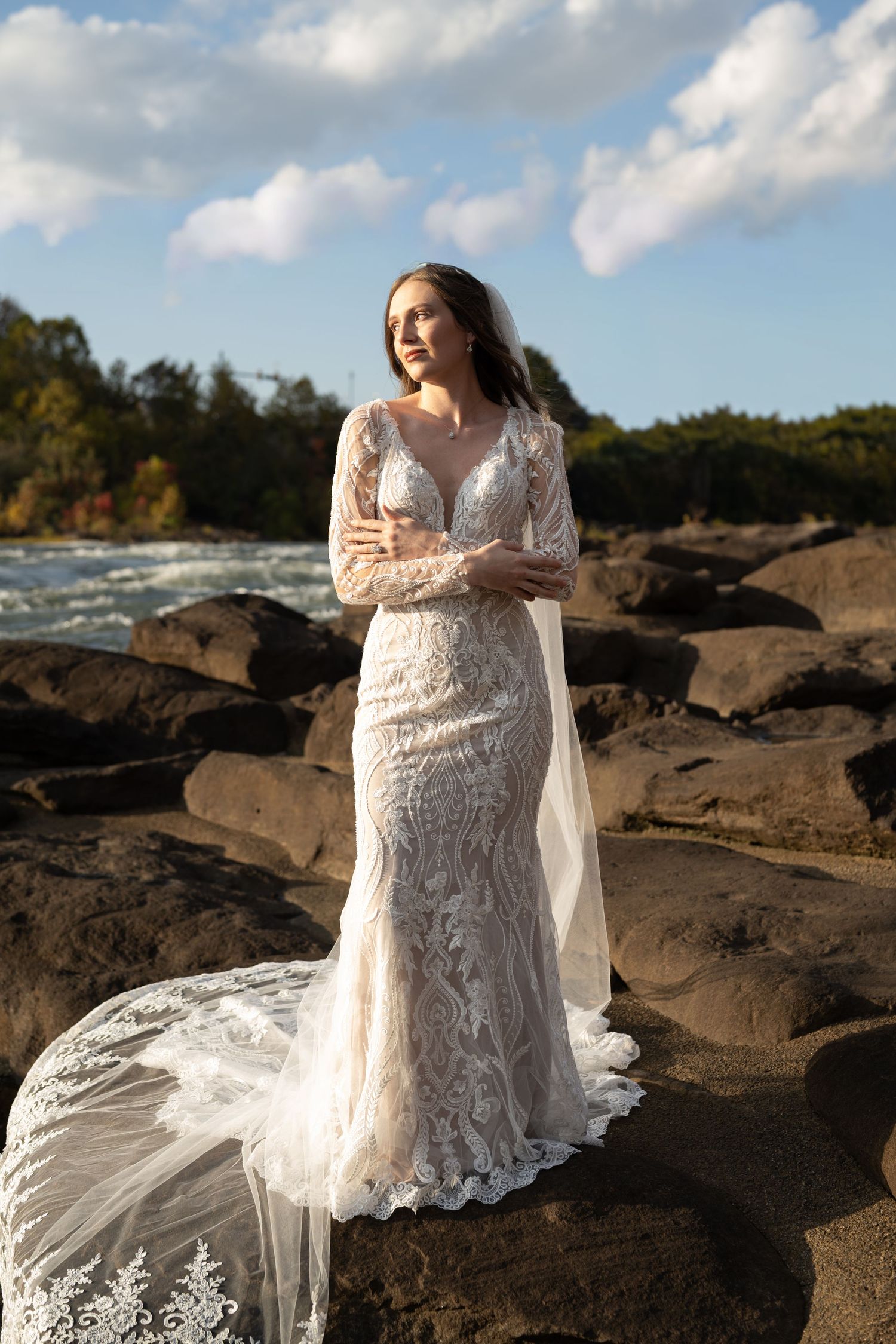 A lace wedding dress with long sleeves photographed against rocky terrain and flowing water on a cloudy day.