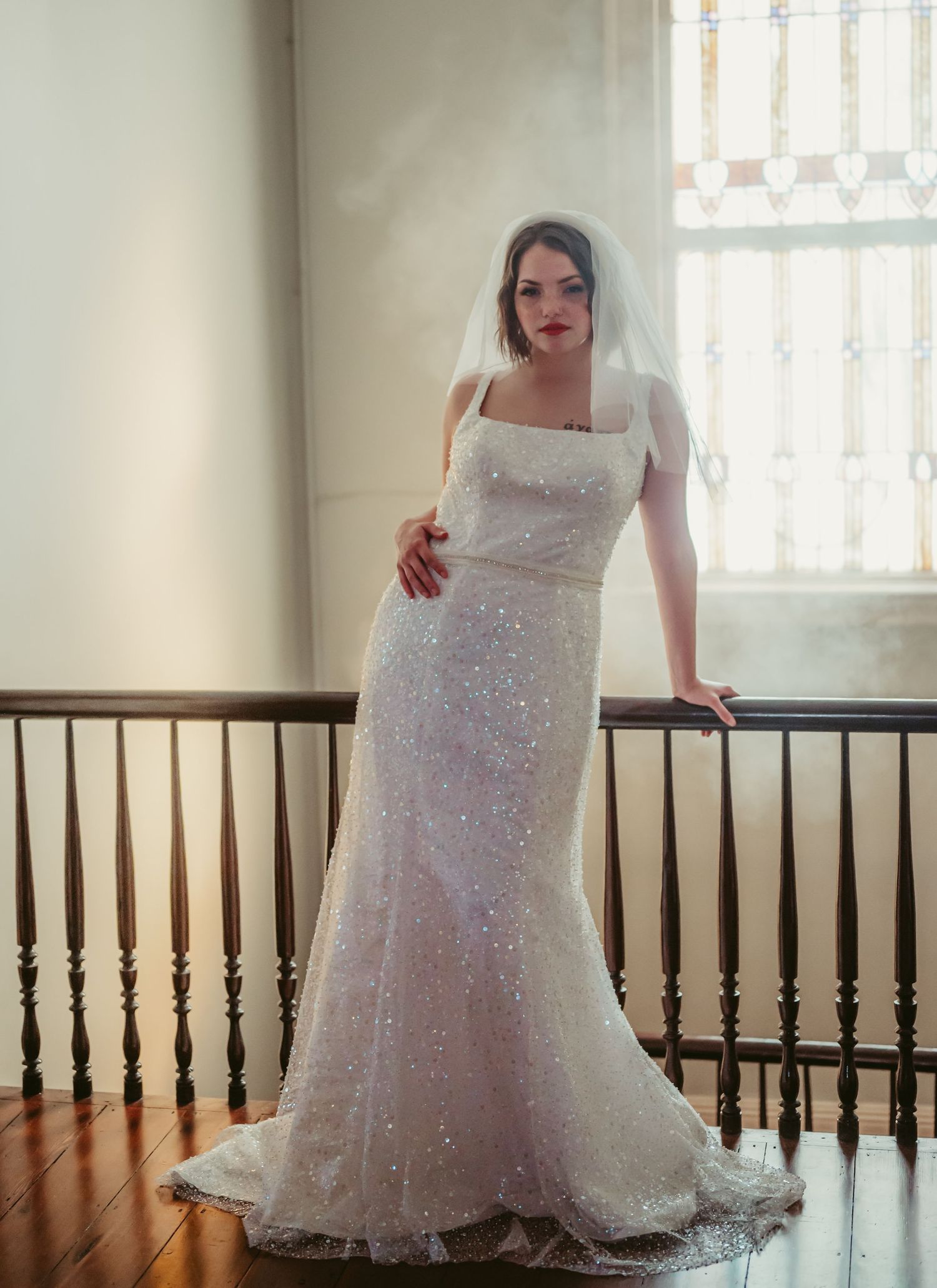 A bride in a sparkling white mermaid wedding gown poses elegantly against a wooden railing in natural window light.
