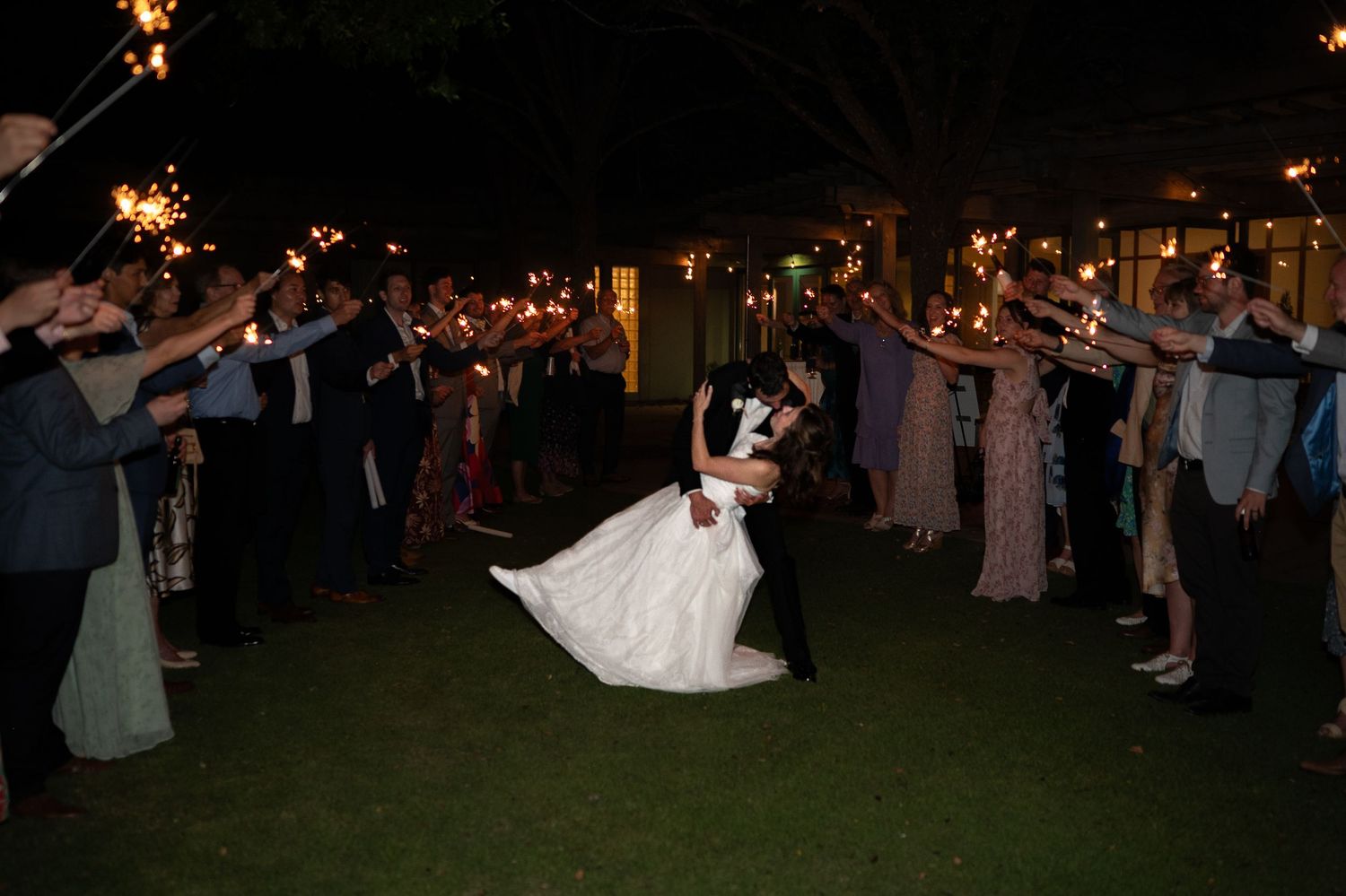 A couple dances together at a nighttime outdoor wedding reception surrounded by sparklers held by guests.